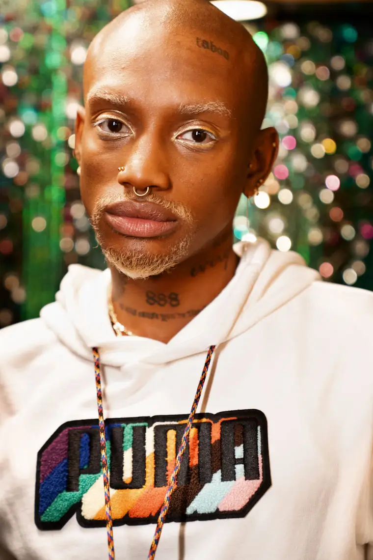 Close-up of a bald person with pierced nose and septum wearing a white hoodie with a colorful PUMA logo against a blurred sparkly background.
