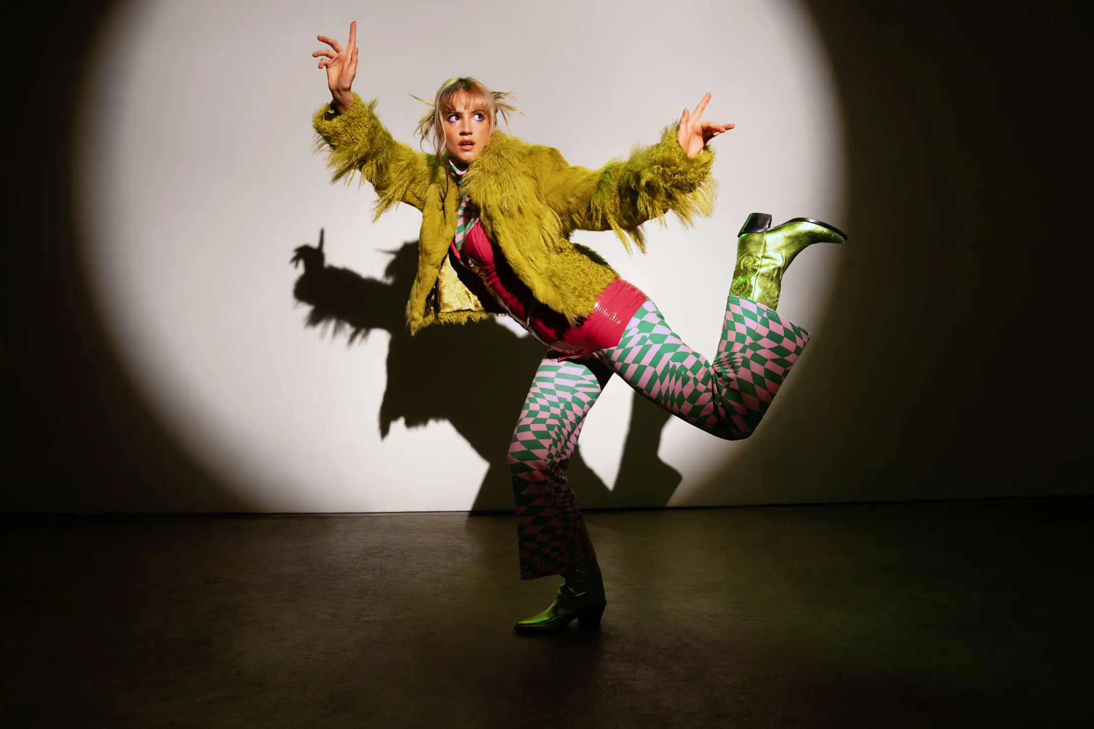 Person in a fuzzy green jacket, pink and green patterned pants, and metallic green boots striking a dynamic pose against a spotlighted wall.