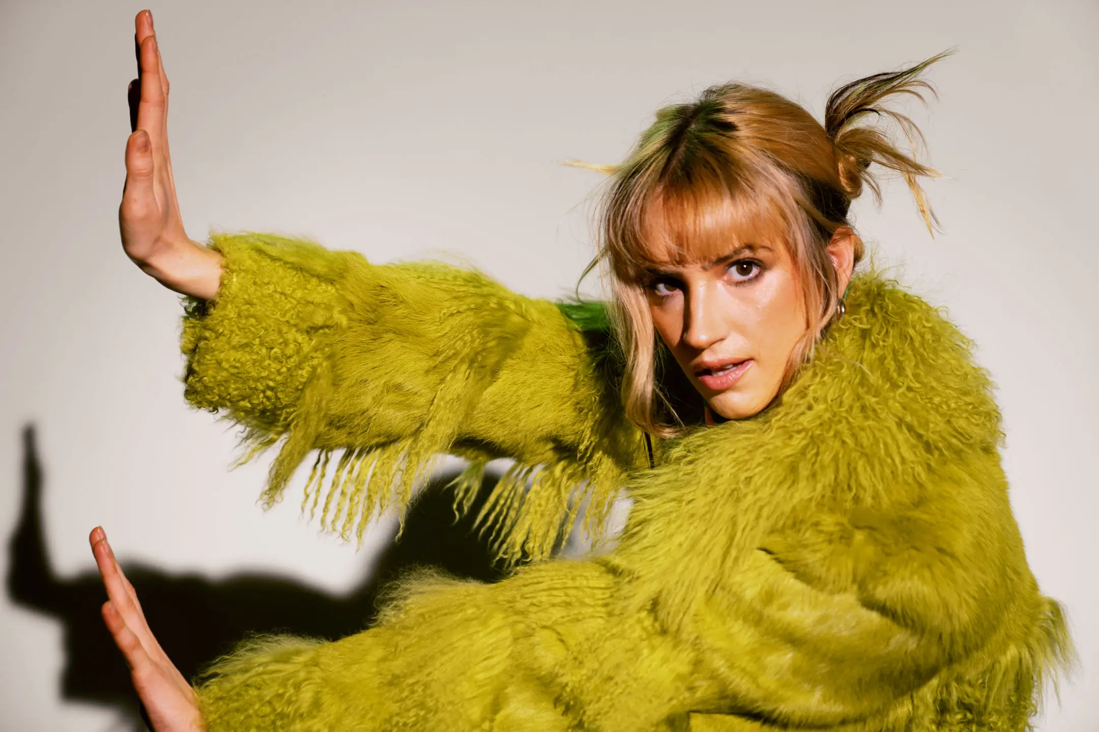 Woman with blonde hair in a green fuzzy jacket posing with both hands raised against a plain background.