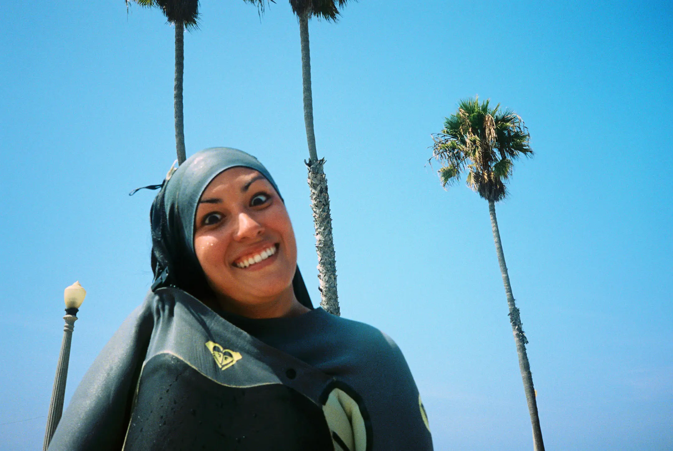 Smiling woman wearing a wetsuit and head covering with palm trees and clear blue sky in the background.