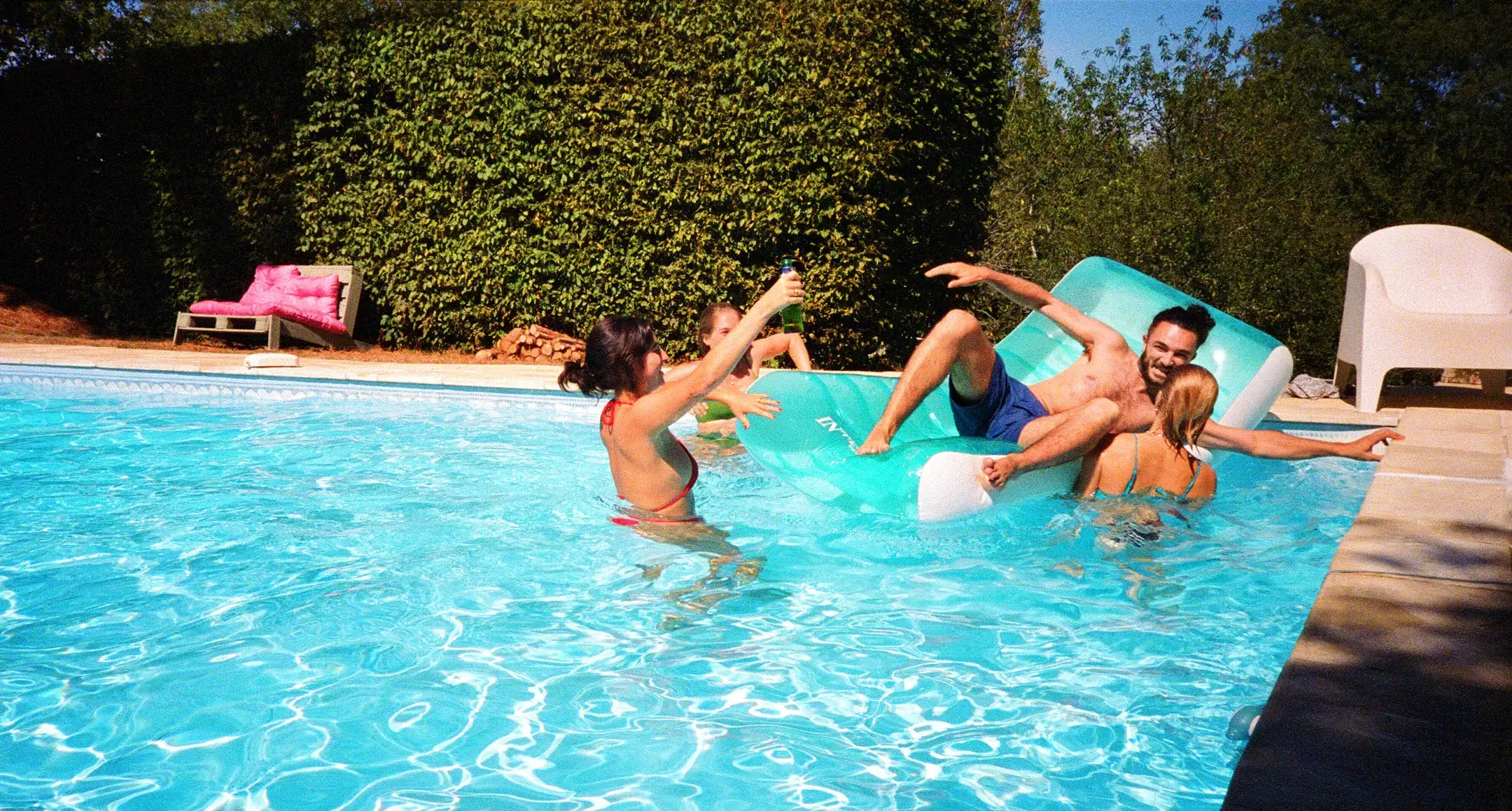 Four people enjoying a sunny day in a pool with a turquoise inflatable float, two holding bottles.