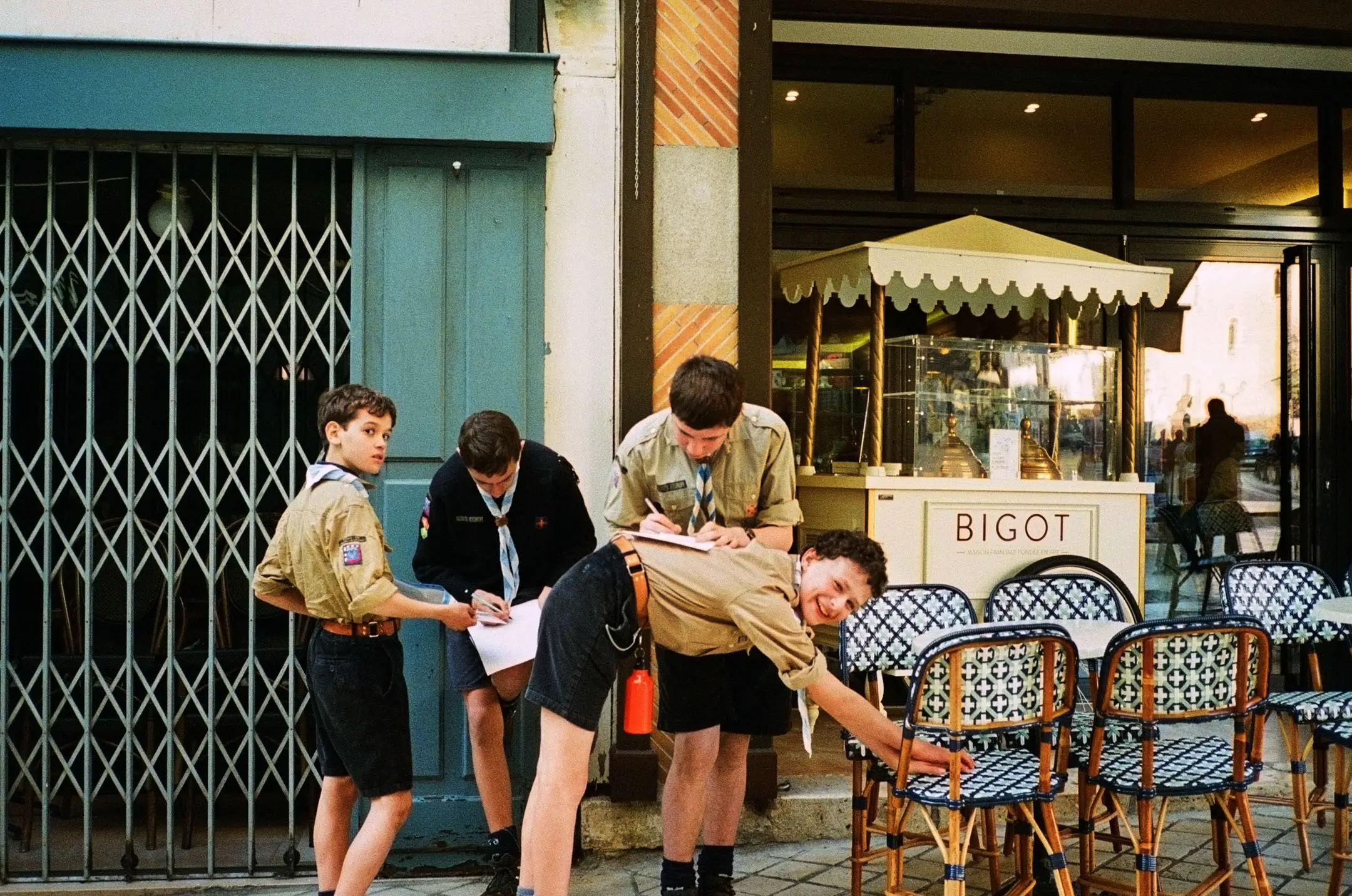 Four Boy Scouts in uniform writing on papers outside a café with patterned chairs and a display labeled BIGOT.