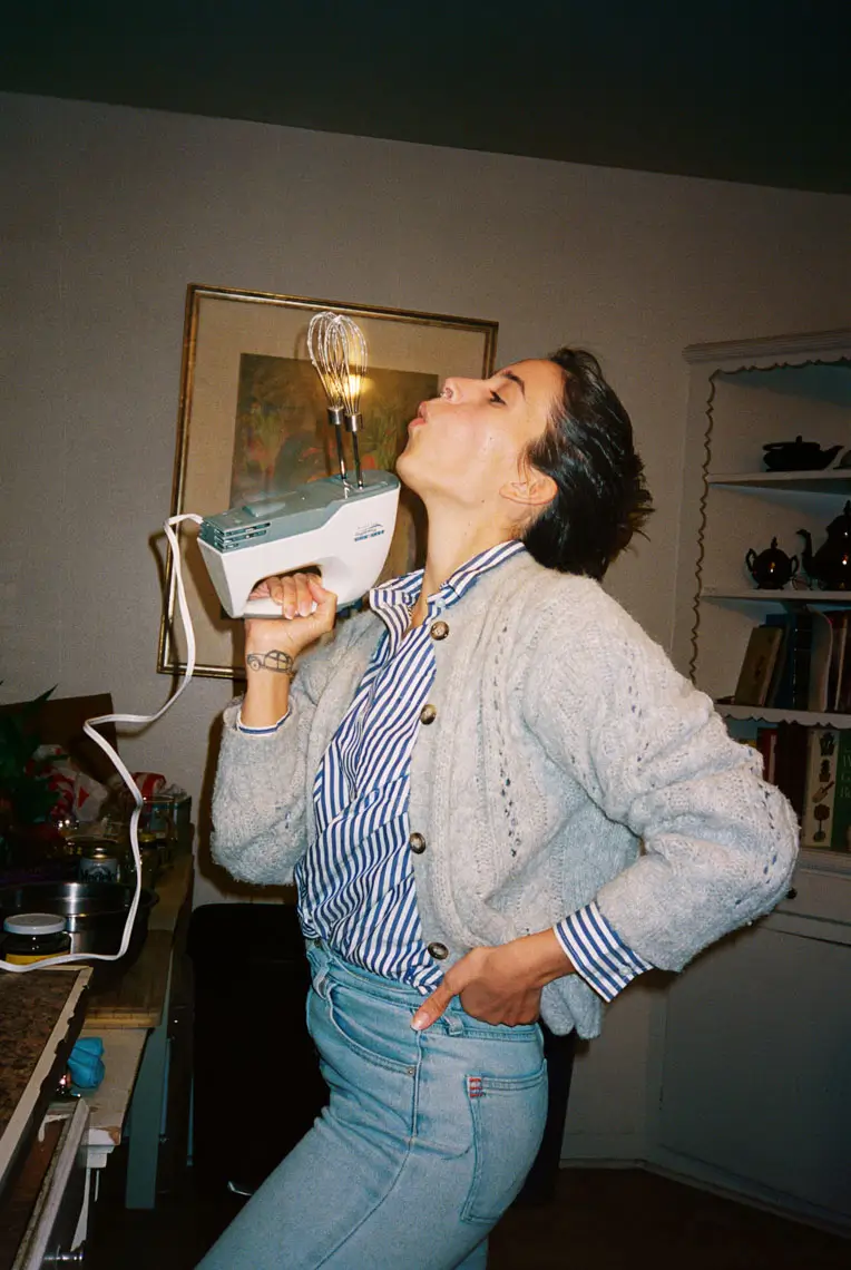Person holding a kitchen hand mixer upright near their face with the beaters lit by a lightbulb, posing playfully in a cozy kitchen.