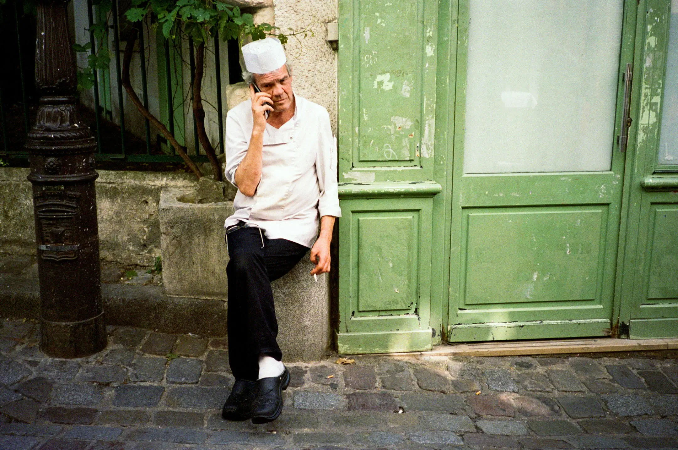 Chef in white uniform and hat sitting on a stone ledge by a green door, smoking and talking on a phone.