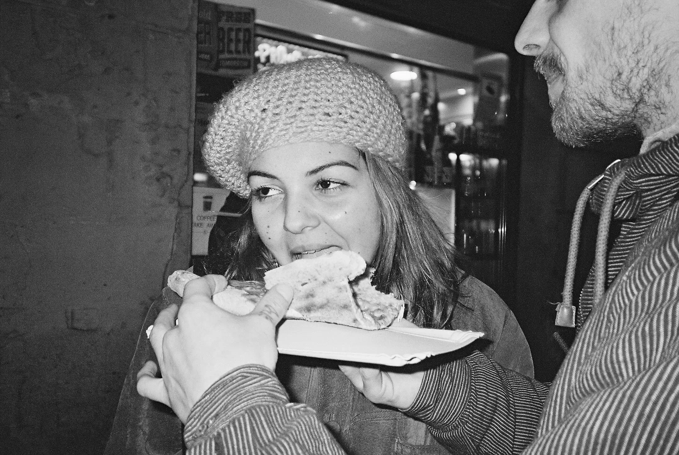 A woman wearing a knitted beret bites into a flatbread sandwich held on a paper plate by a man with a beard in a striped hoodie.