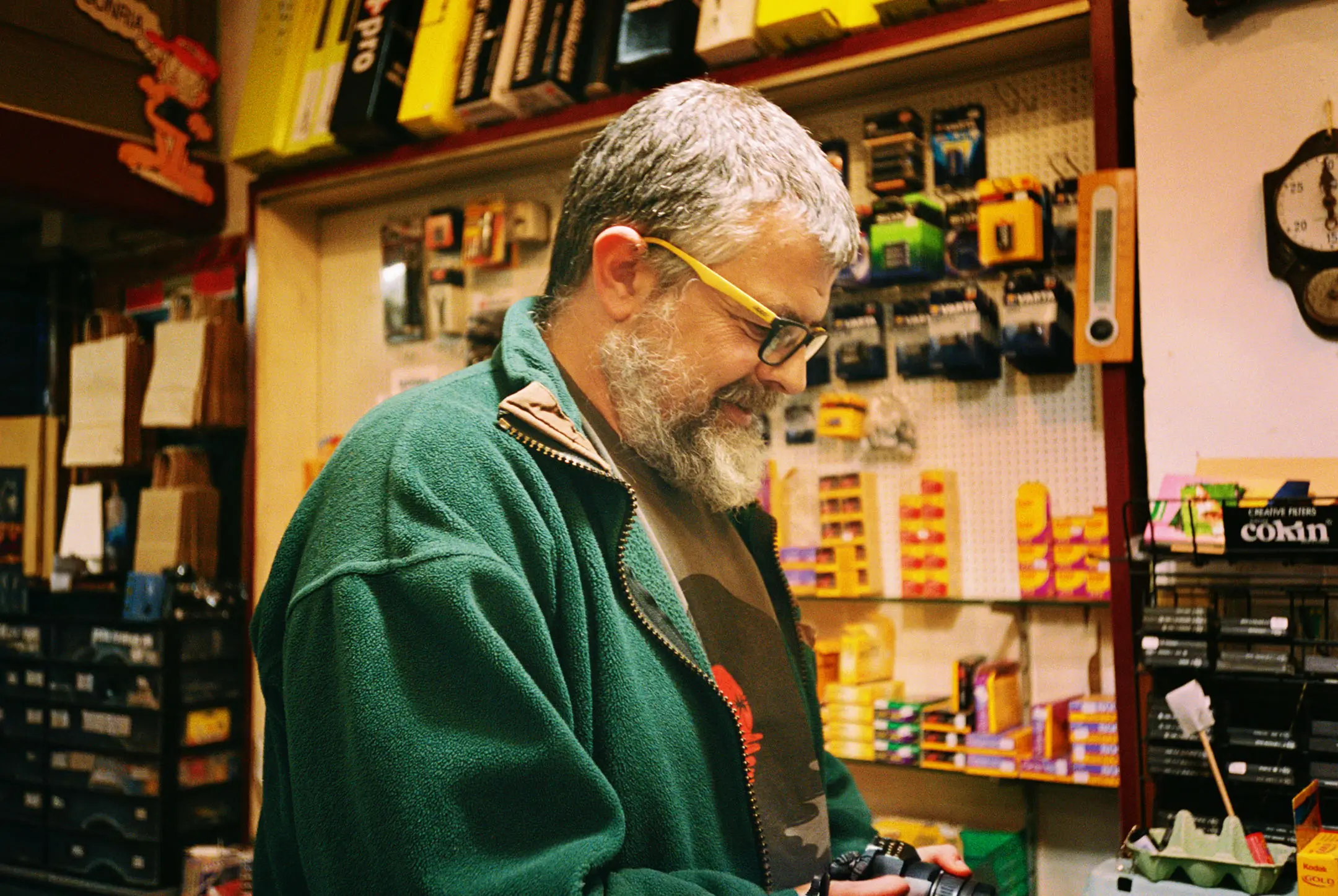 Smiling man with gray hair and beard wearing glasses and green jacket holding a camera in a shop filled with photography supplies.