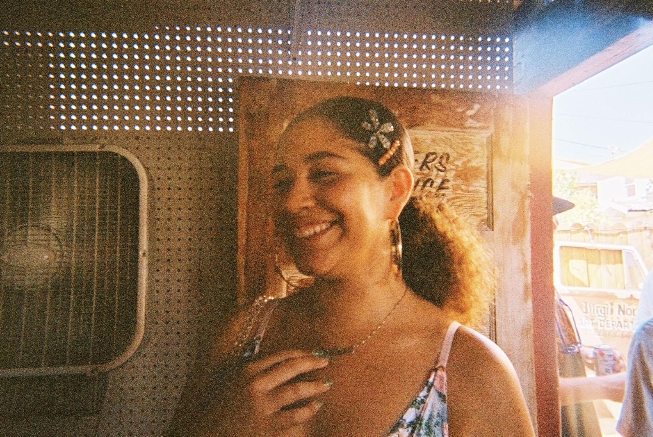 Smiling woman with curly hair in a ponytail, wearing hoop earrings and floral dress, standing indoors near a wooden door with sunlight shining in.
