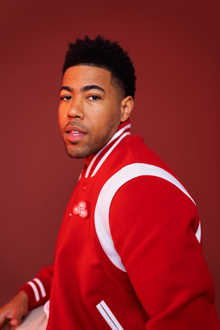 Man with short curly hair wearing a red and white varsity jacket against a red background.