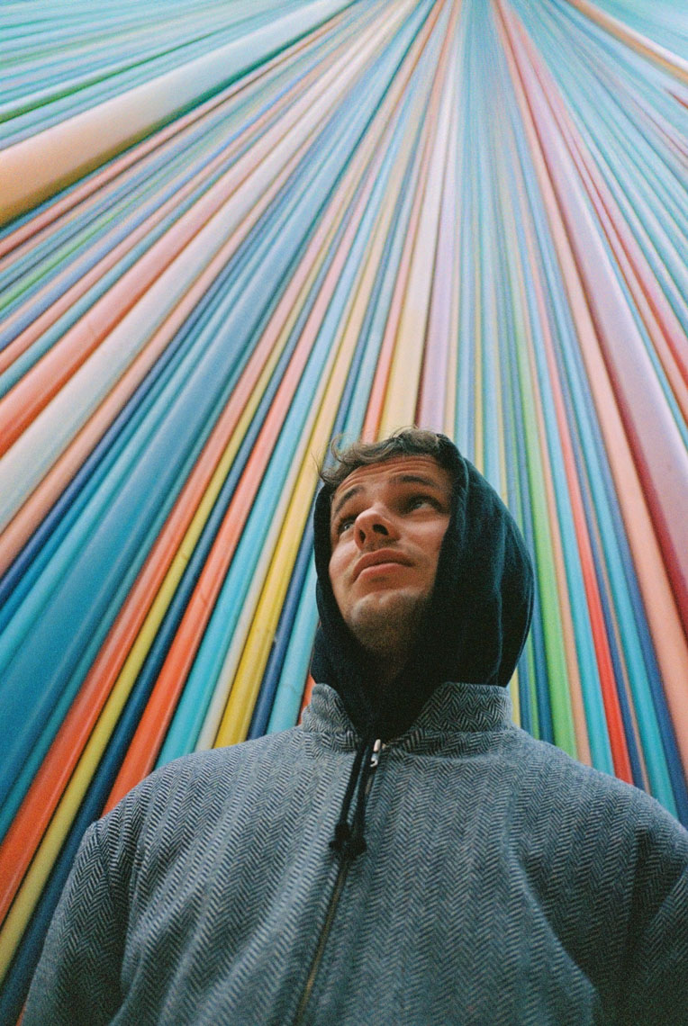Young man in a hooded jacket looking up against a vibrant backdrop of vertical colorful lines.