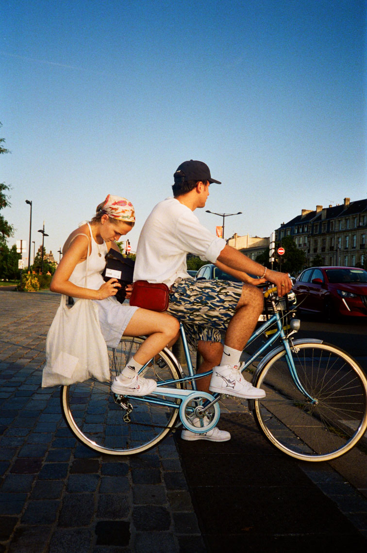 Man and woman riding a blue bicycle on a city street during golden hour; woman holds a camera and wears a headscarf, man wears a cap and camouflage shorts.