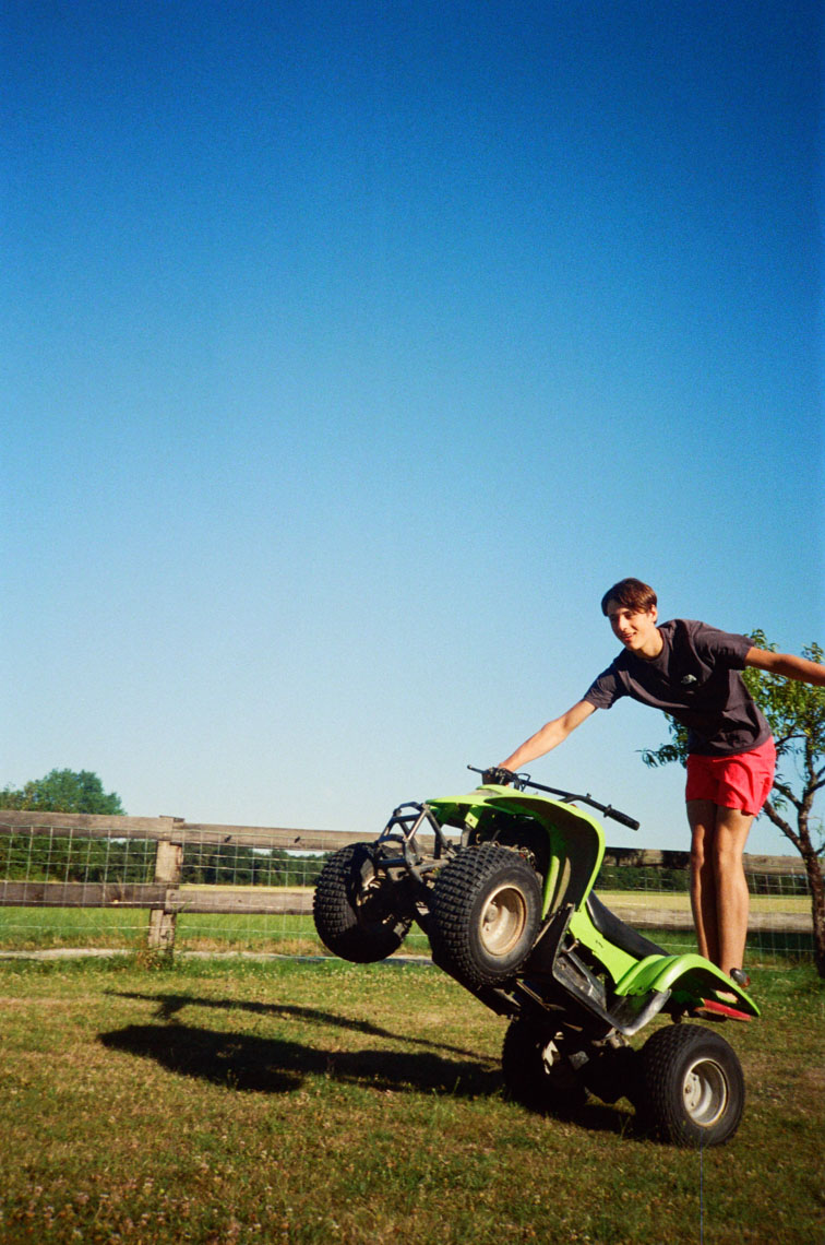 Teenage boy balancing on the rear wheels of a green ATV on a grassy lawn near a wooden fence.