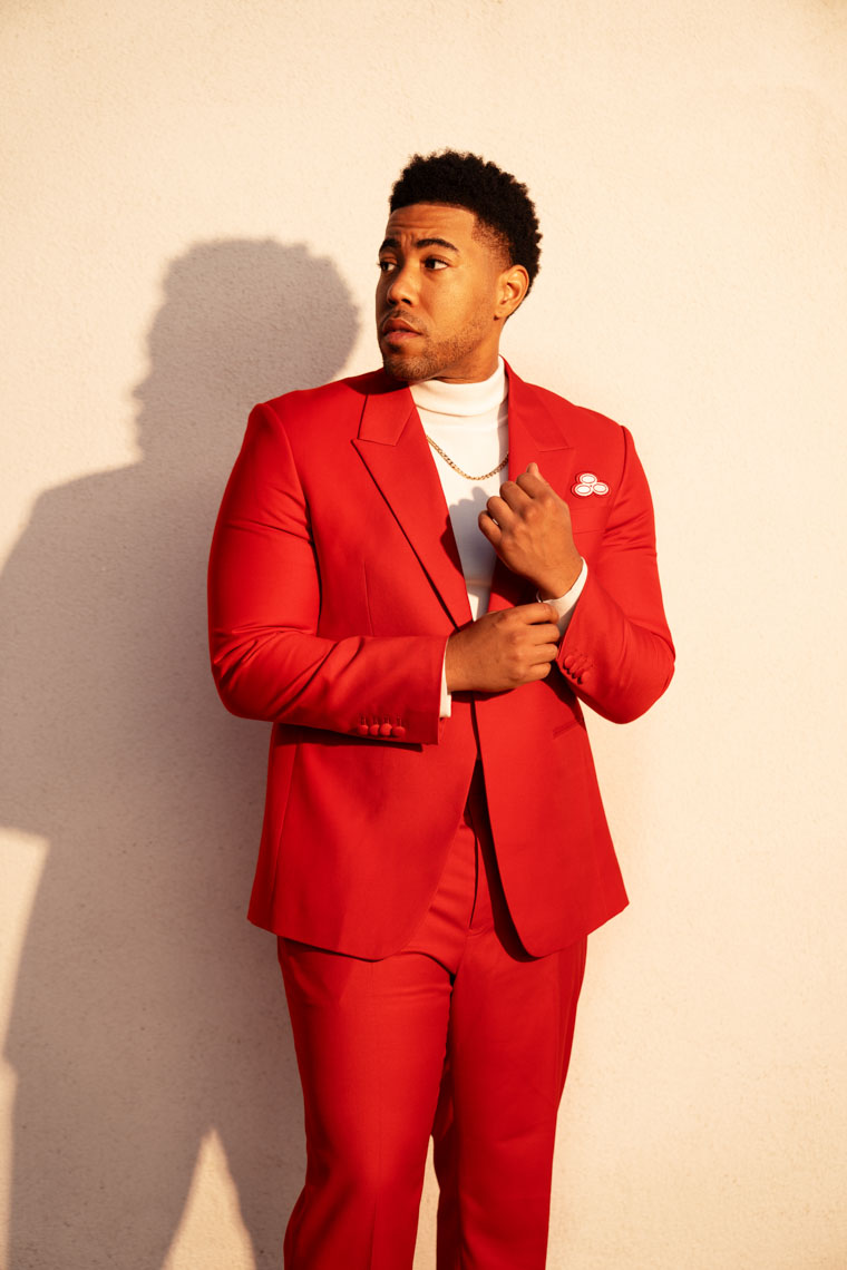 Man wearing a bright red suit and white turtleneck standing against a beige wall, adjusting his cuff with shadow cast behind him.