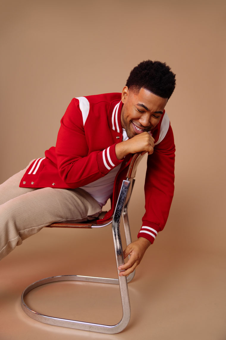 Smiling man in a red varsity jacket leaning and holding the backrest of a modern metal and leather chair against a beige background.