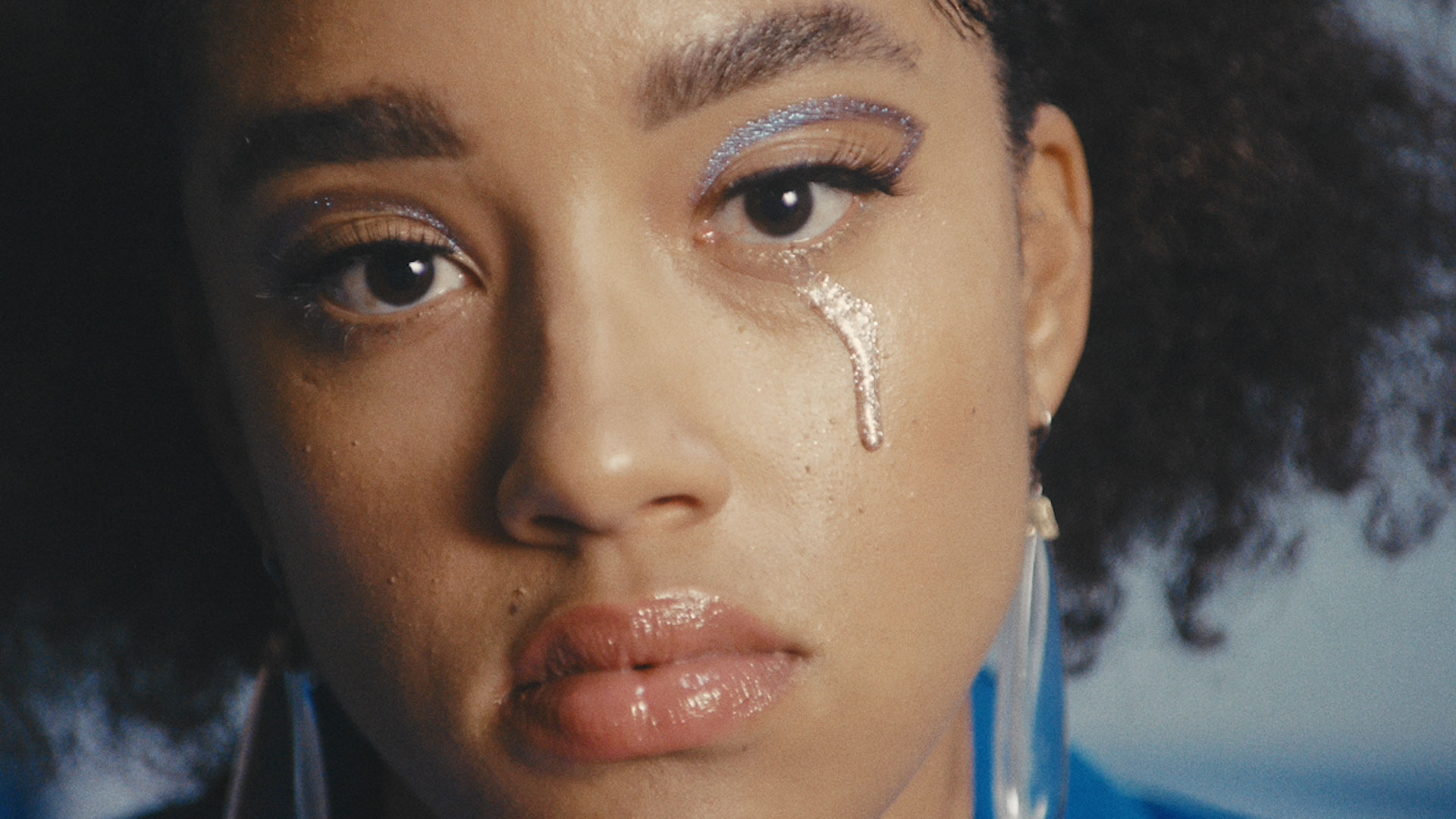 Close-up of a young woman with glittery blue eyeshadow and a silver glitter tear streak on her cheek.