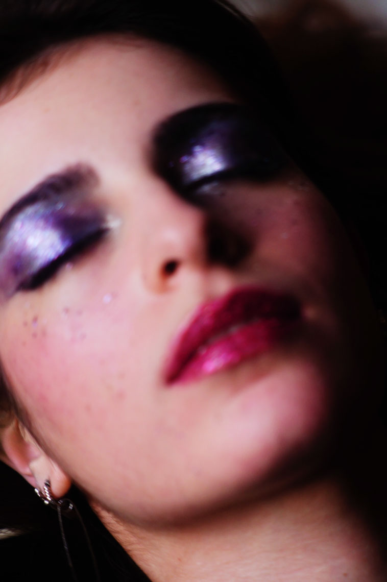 Close-up of a person with purple glitter eyeshadow, pink glossy lips, and tiny face glitter, eyes closed.