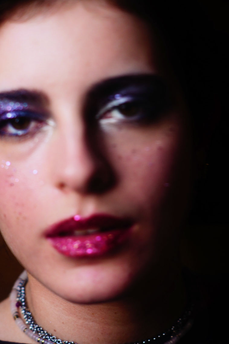 Close-up portrait of a person with glittery purple eyeshadow, pink glitter lipstick, and a beaded necklace.