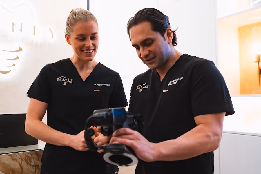 Two medical professionals in black scrubs looking at a handheld device together and smiling.