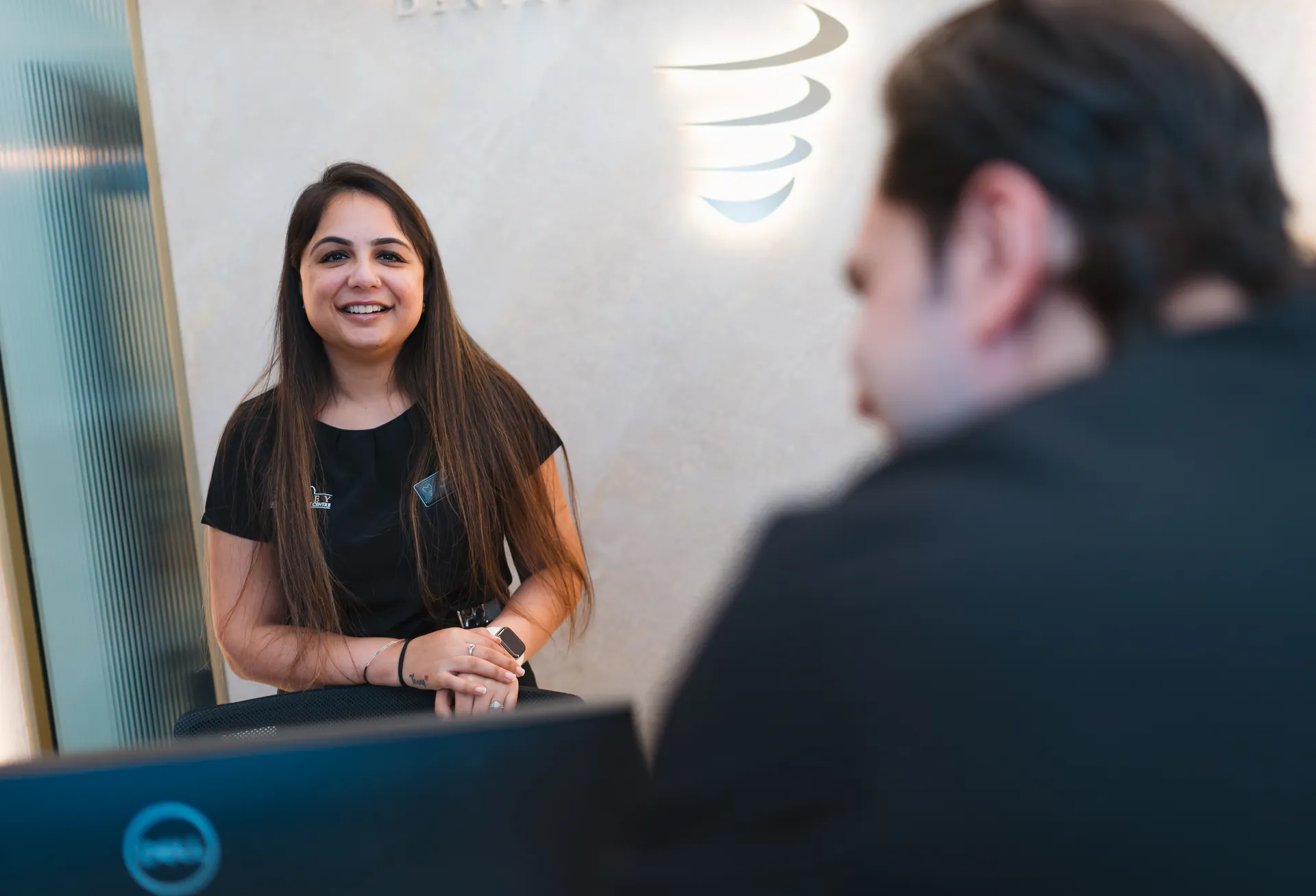 Smiling woman with long hair leaning on a chair, talking to a man seen from behind in an office setting.