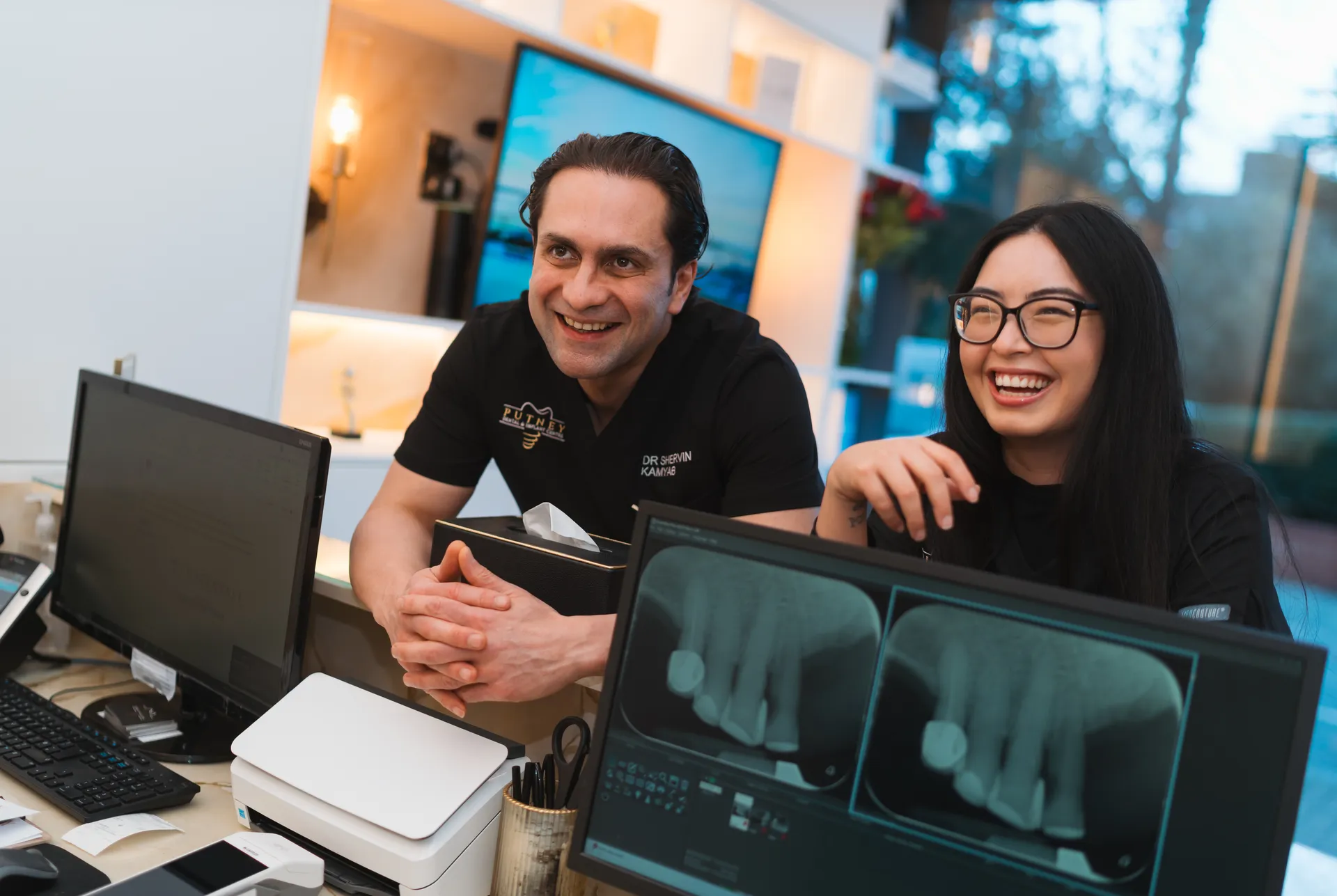 Two dental professionals smiling and leaning over a desk with computer monitors displaying dental X-rays.