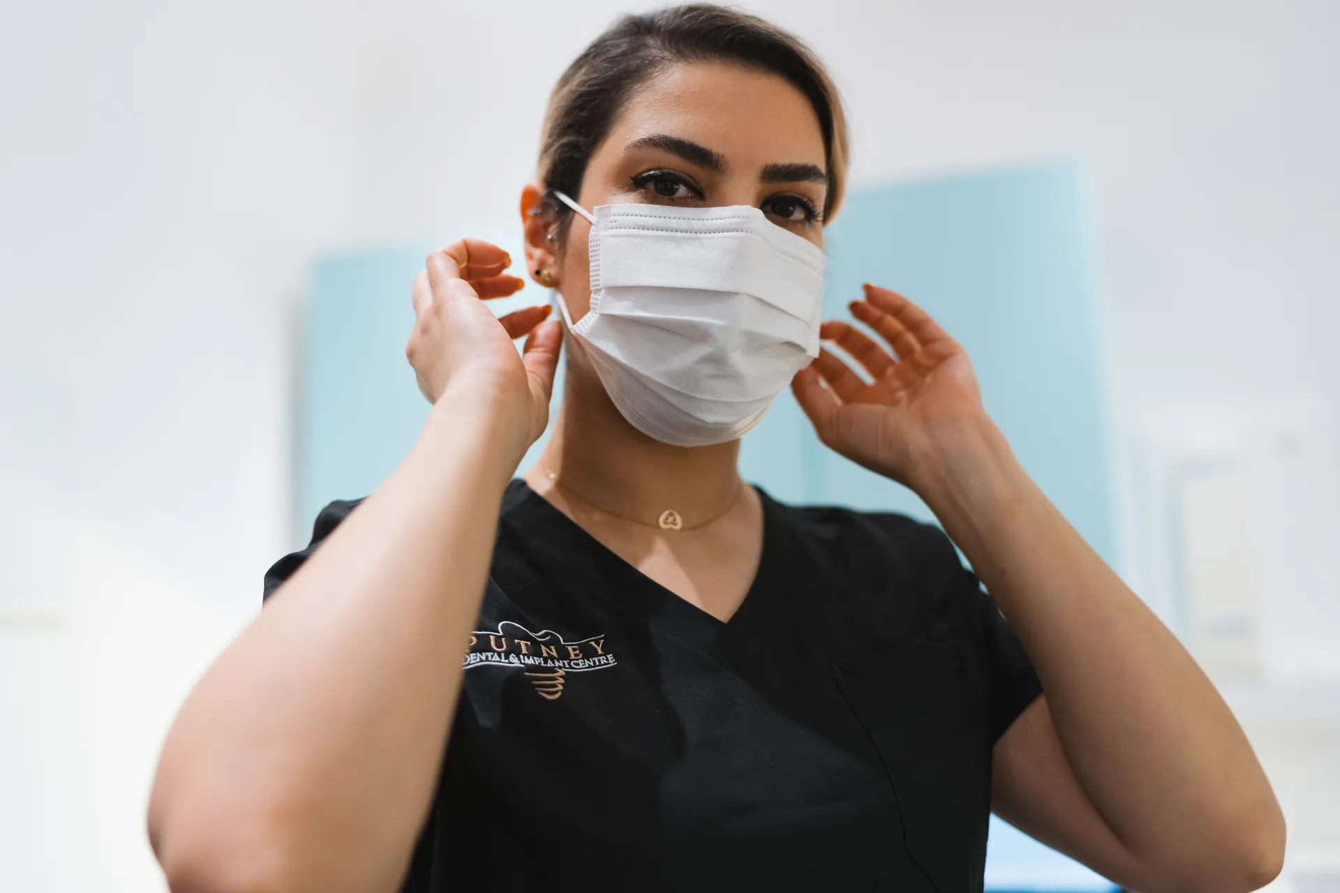 Woman wearing a white medical face mask and black scrubs adjusting the mask straps near her ears.
