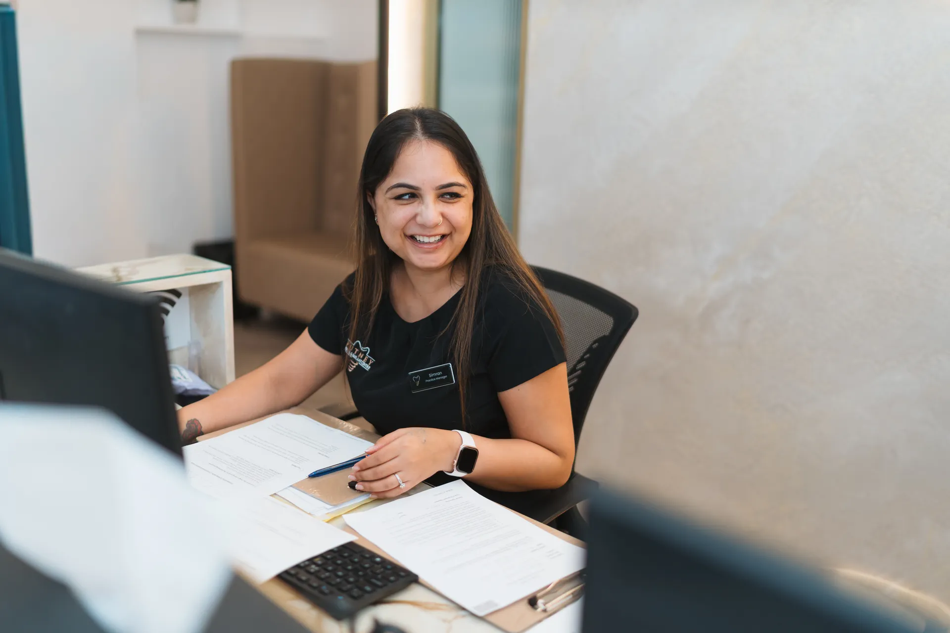 Smiling woman with long dark hair sitting at a desk with documents, a pen, and a computer, wearing a black shirt and a smartwatch.