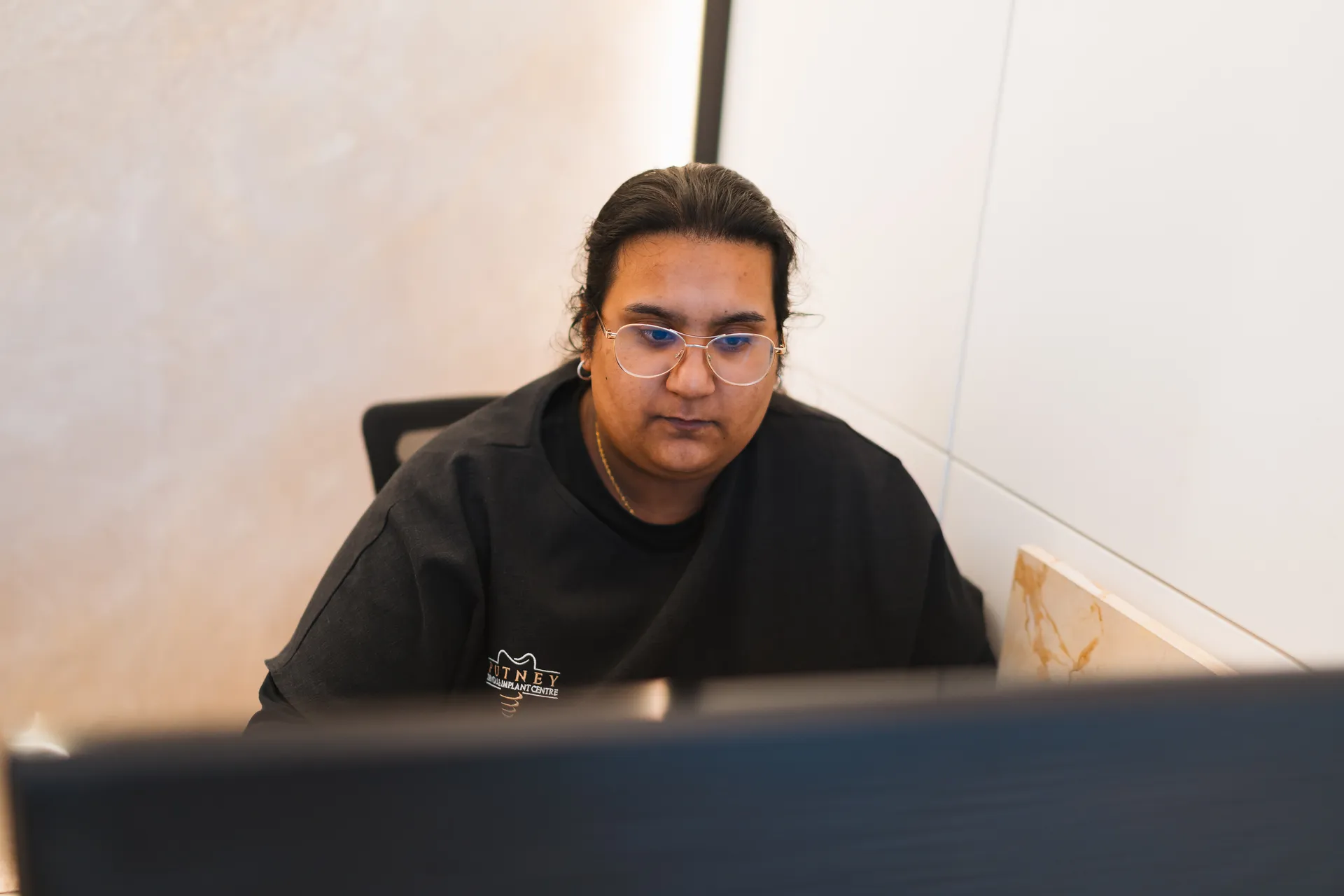 Person with glasses and dark hair tied back, focused on a computer screen in an office setting.