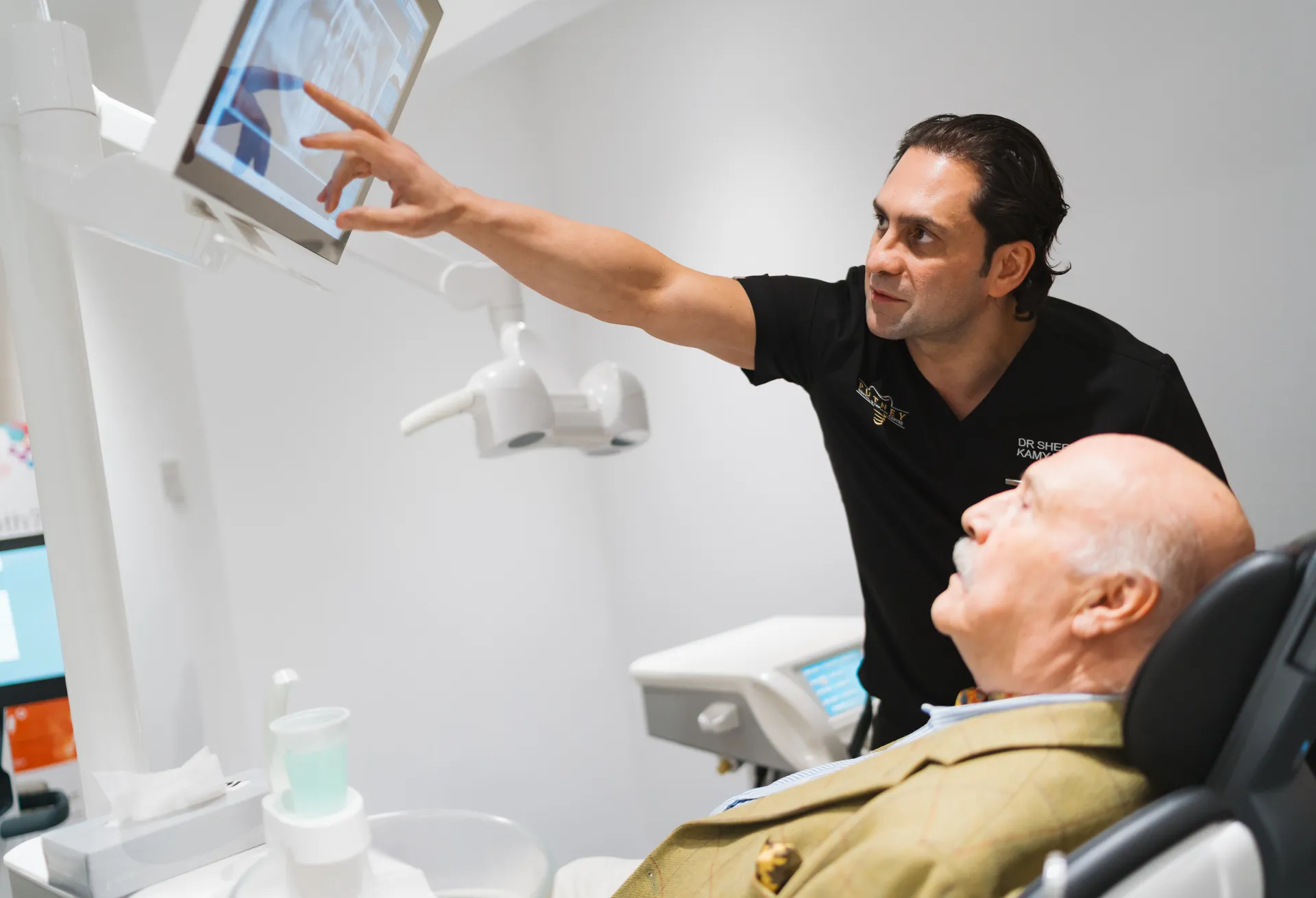 Dentist pointing at an X-ray on a monitor while an elderly male patient sits in the dental chair looking at the screen.