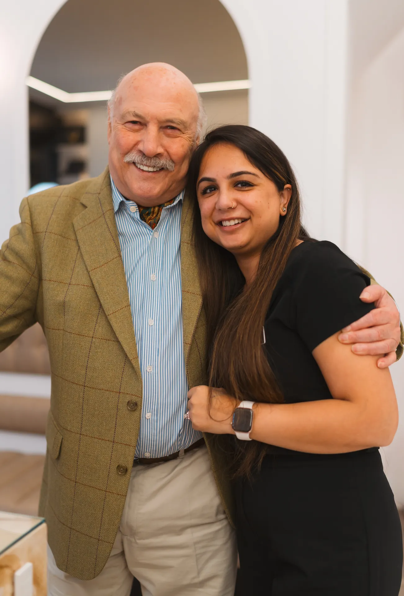 Smiling elderly man in a green plaid jacket hugging a smiling young woman with long hair and a smartwatch.