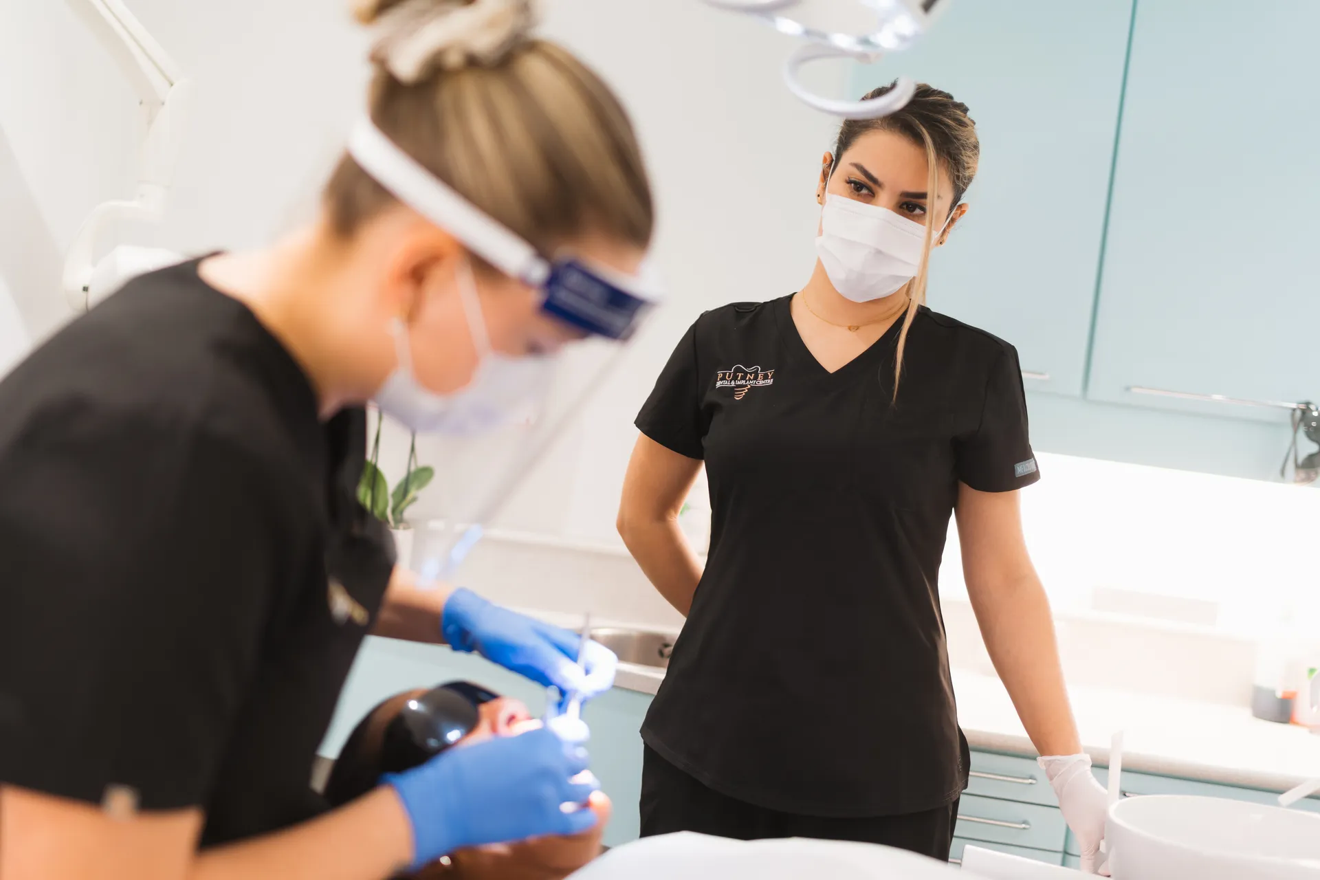 Female dentist in blue gloves treating a patient while a dental assistant in black scrubs and mask observes.