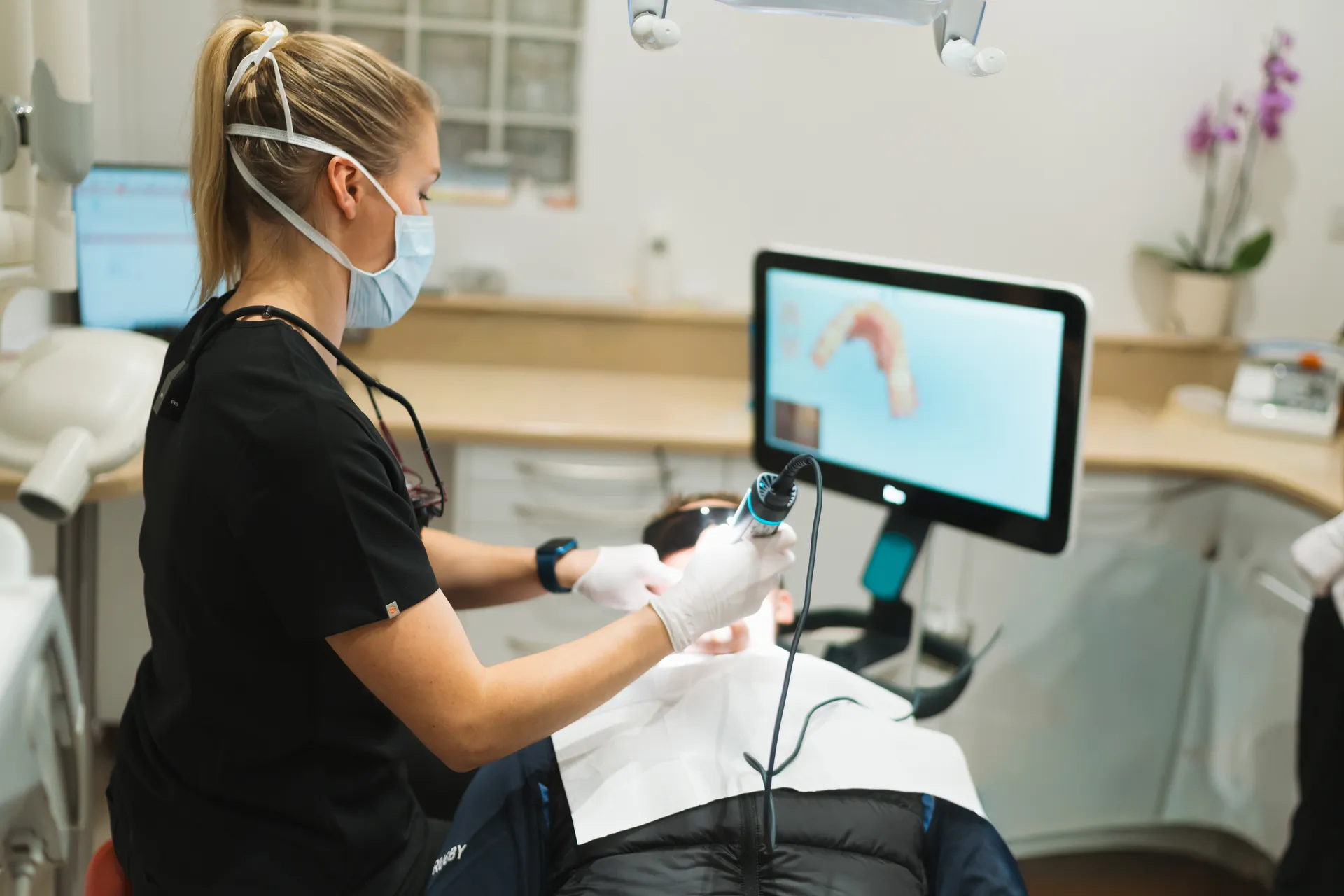 Dental professional wearing a mask and gloves uses a handheld scanner on a patient reclining in a dental chair with a digital dental model displayed on a monitor.