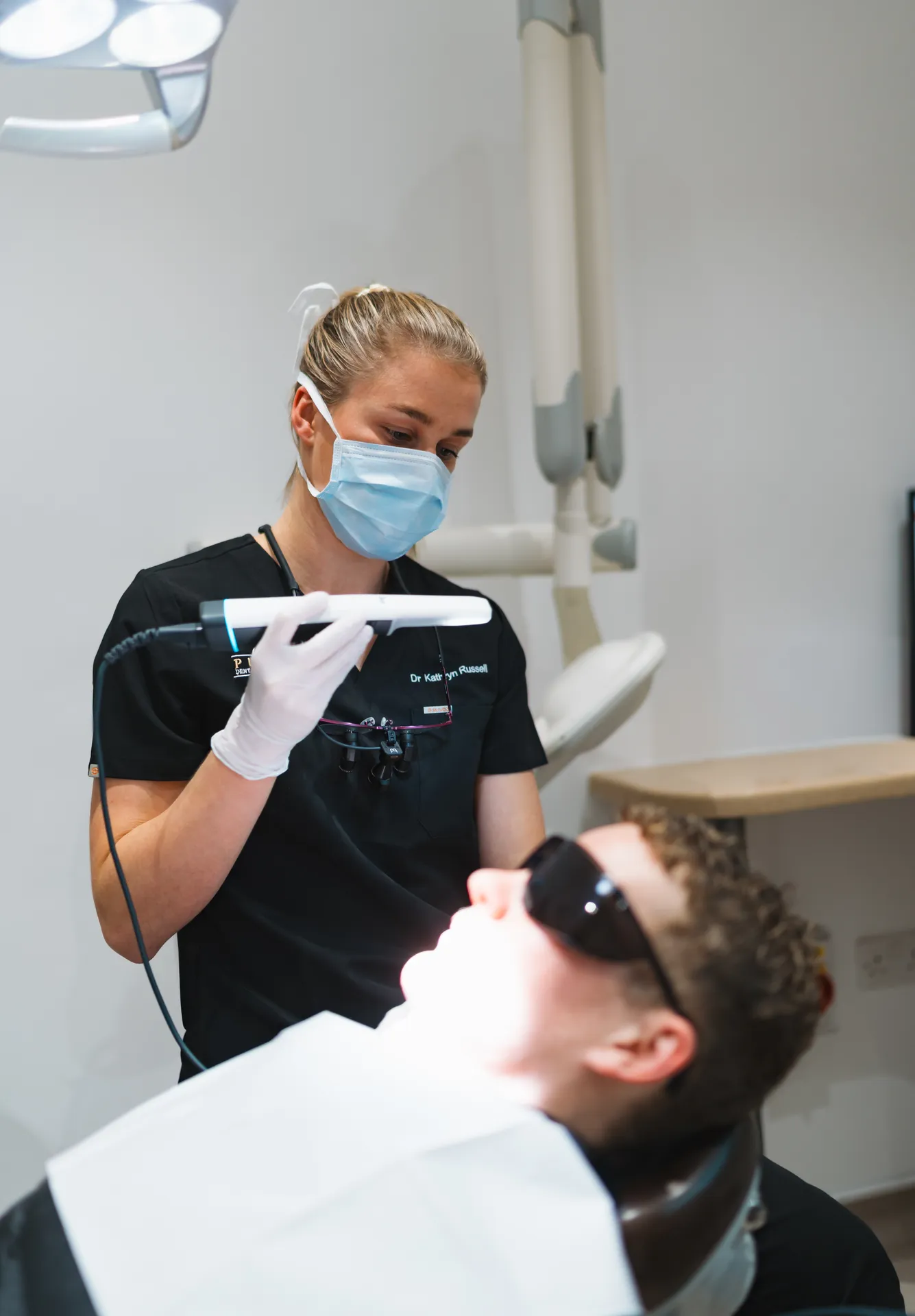 Dentist wearing mask and gloves using a handheld device to examine a patient reclining in a dental chair with protective glasses.