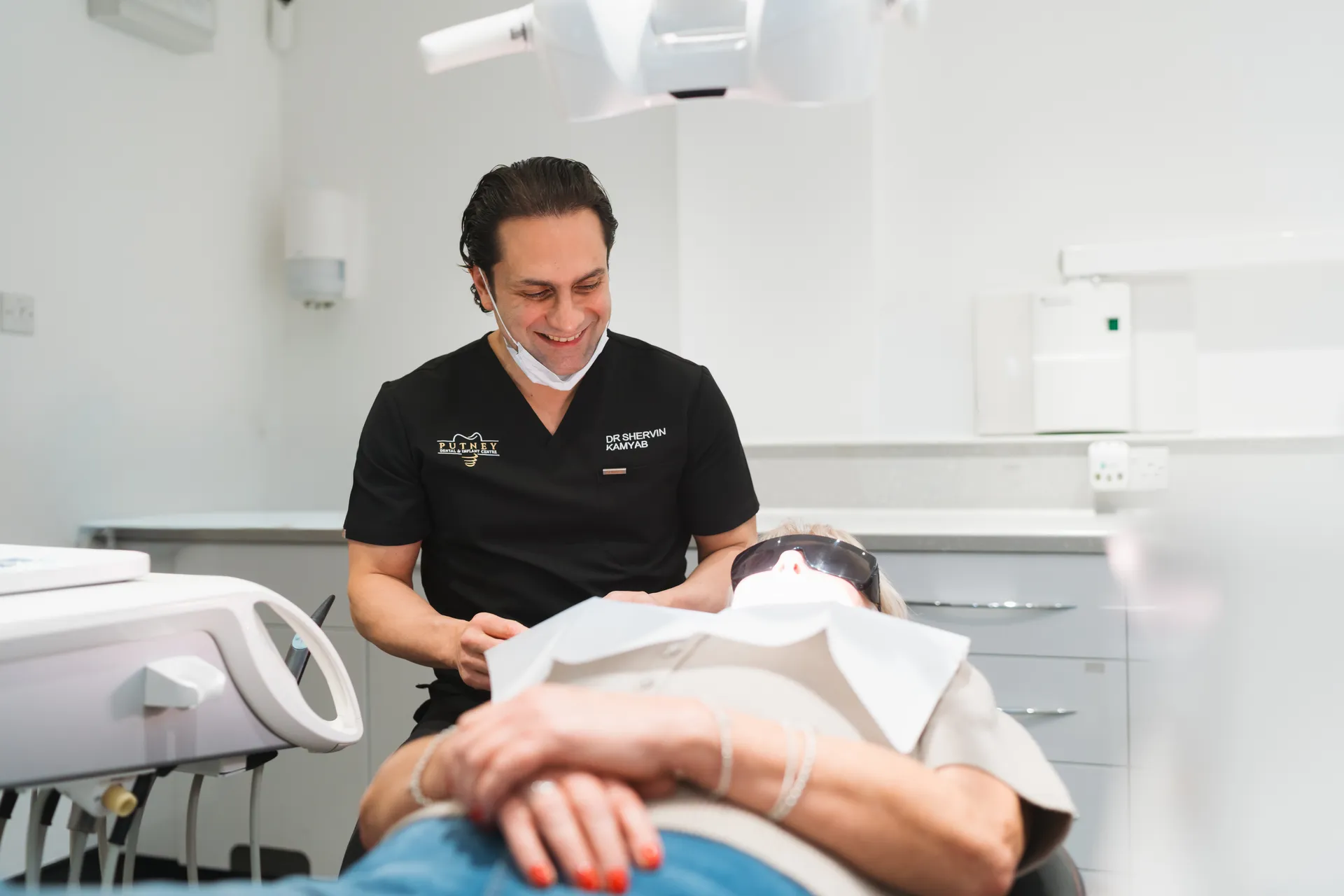 Dentist in black scrubs smiling and preparing to treat a patient lying on a dental chair wearing protective glasses.