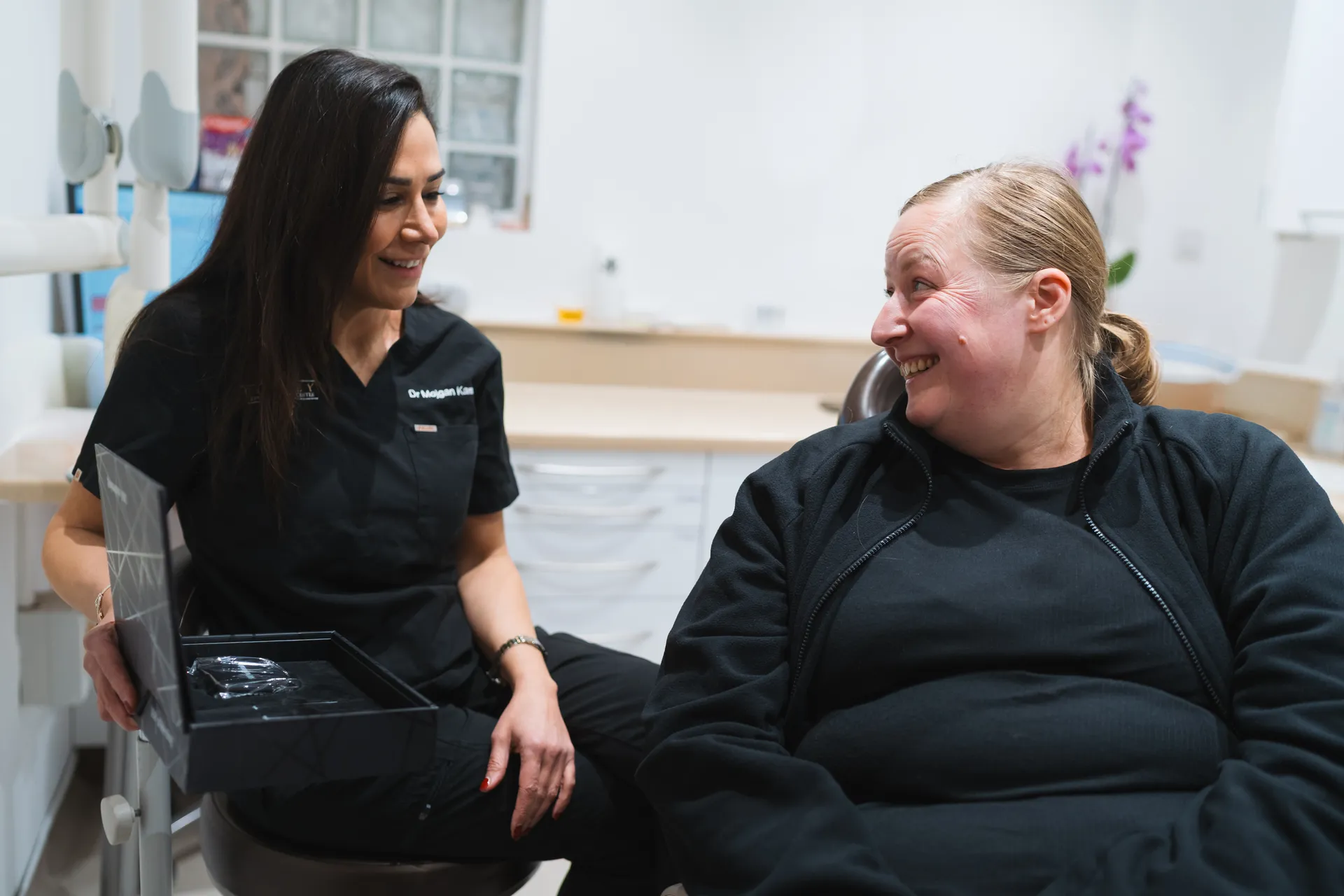 Female dental professional smiling and showing a clear aligner in a box to a smiling patient seated in a dental office.