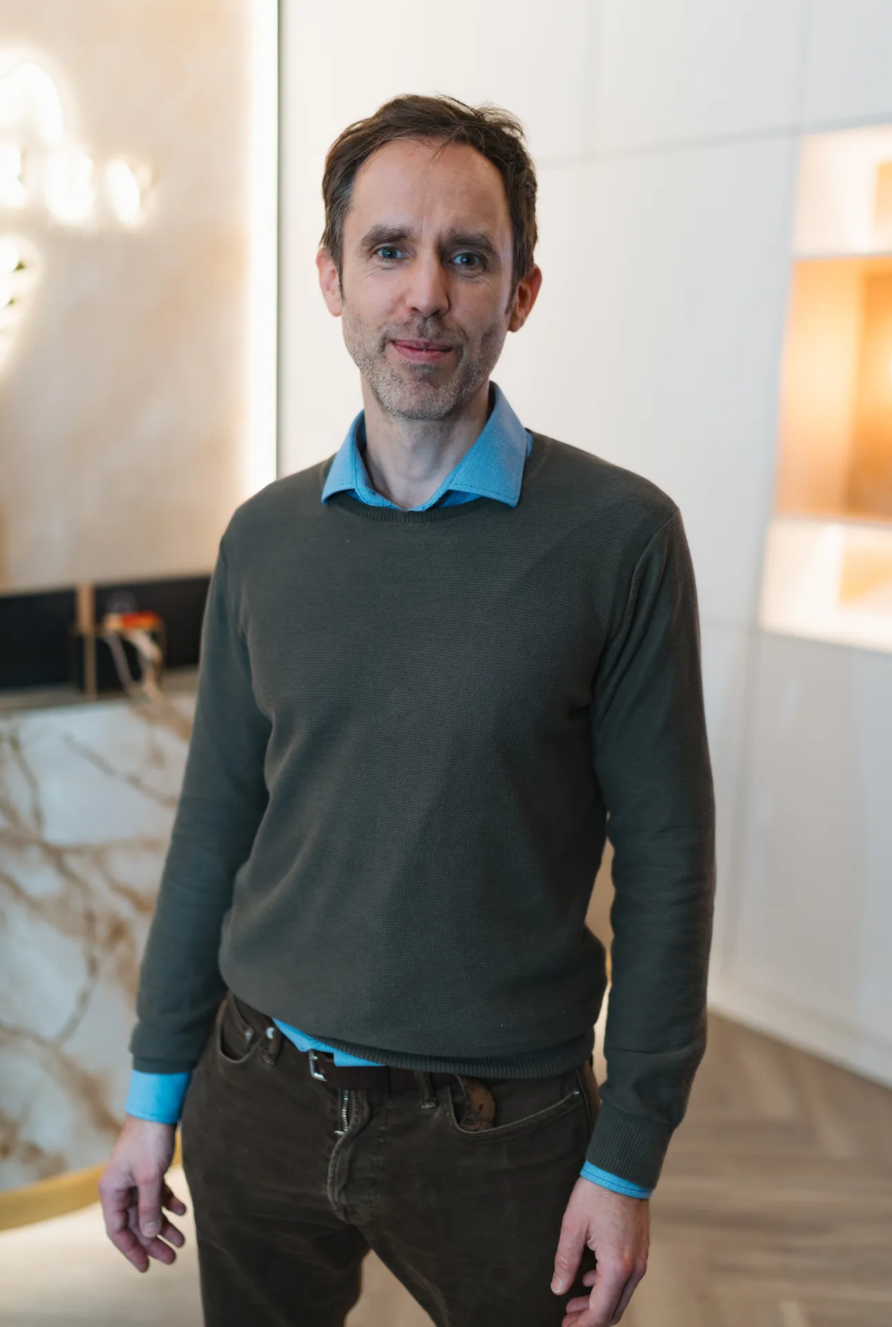 Man with short dark hair and light beard wearing a blue collared shirt under a dark green sweater standing indoors.
