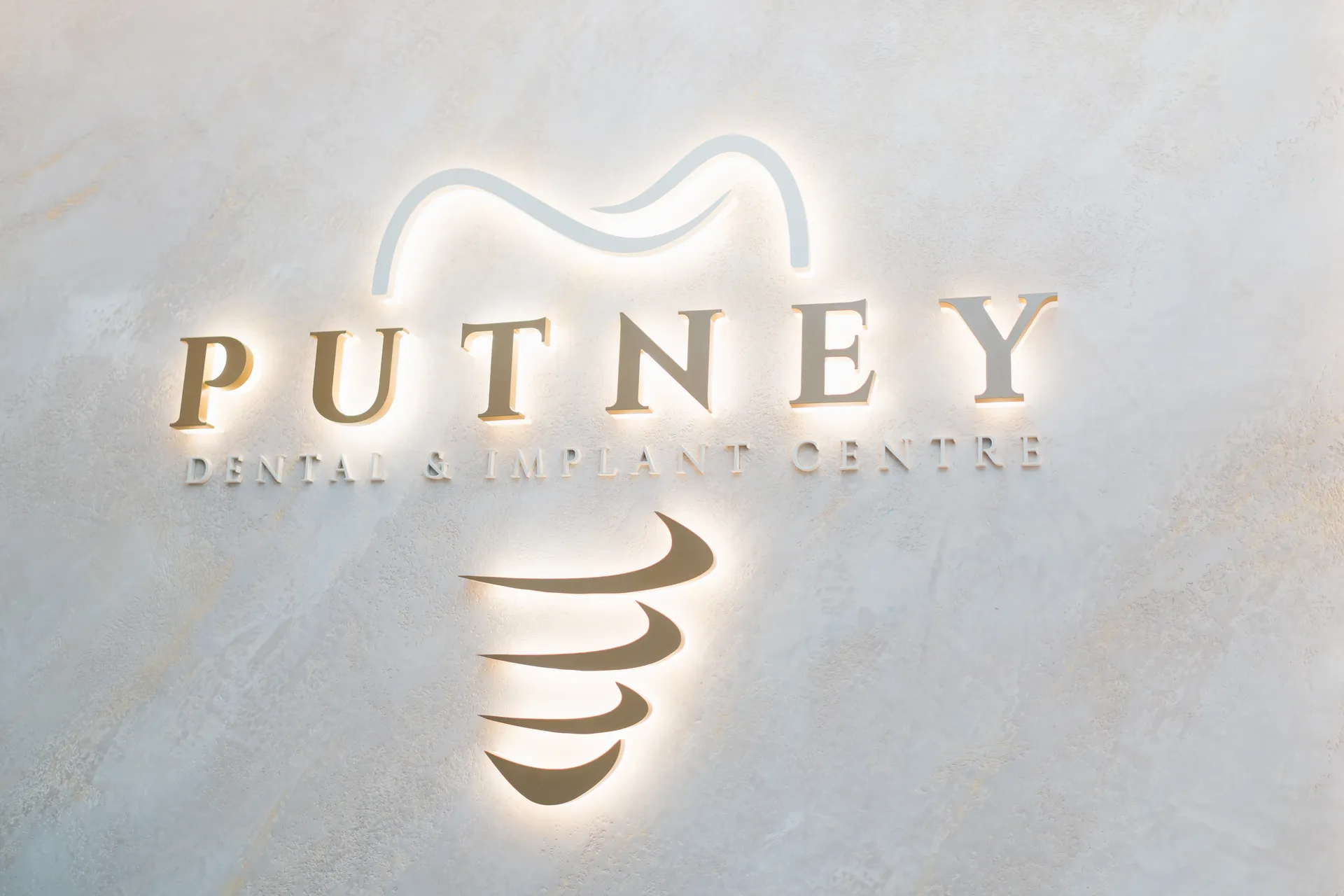 Illuminated sign for Putney Dental & Implant Centre featuring stylized tooth and dental implant design on textured wall.