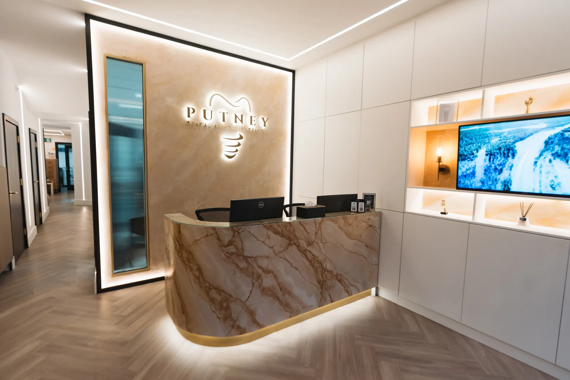 Modern dental clinic reception with a marble desk, illuminated PUTNEY Dental & Implant Clinic logo, and a wall-mounted TV showing a nature scene.