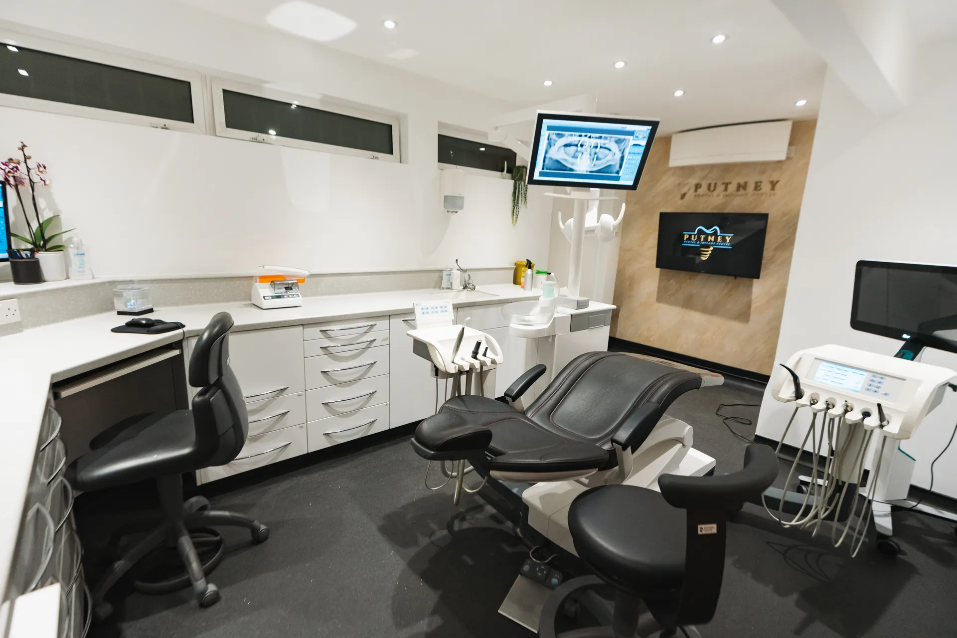 Modern dental examination room with black patient chair, dental instruments, and monitors displaying X-ray images.