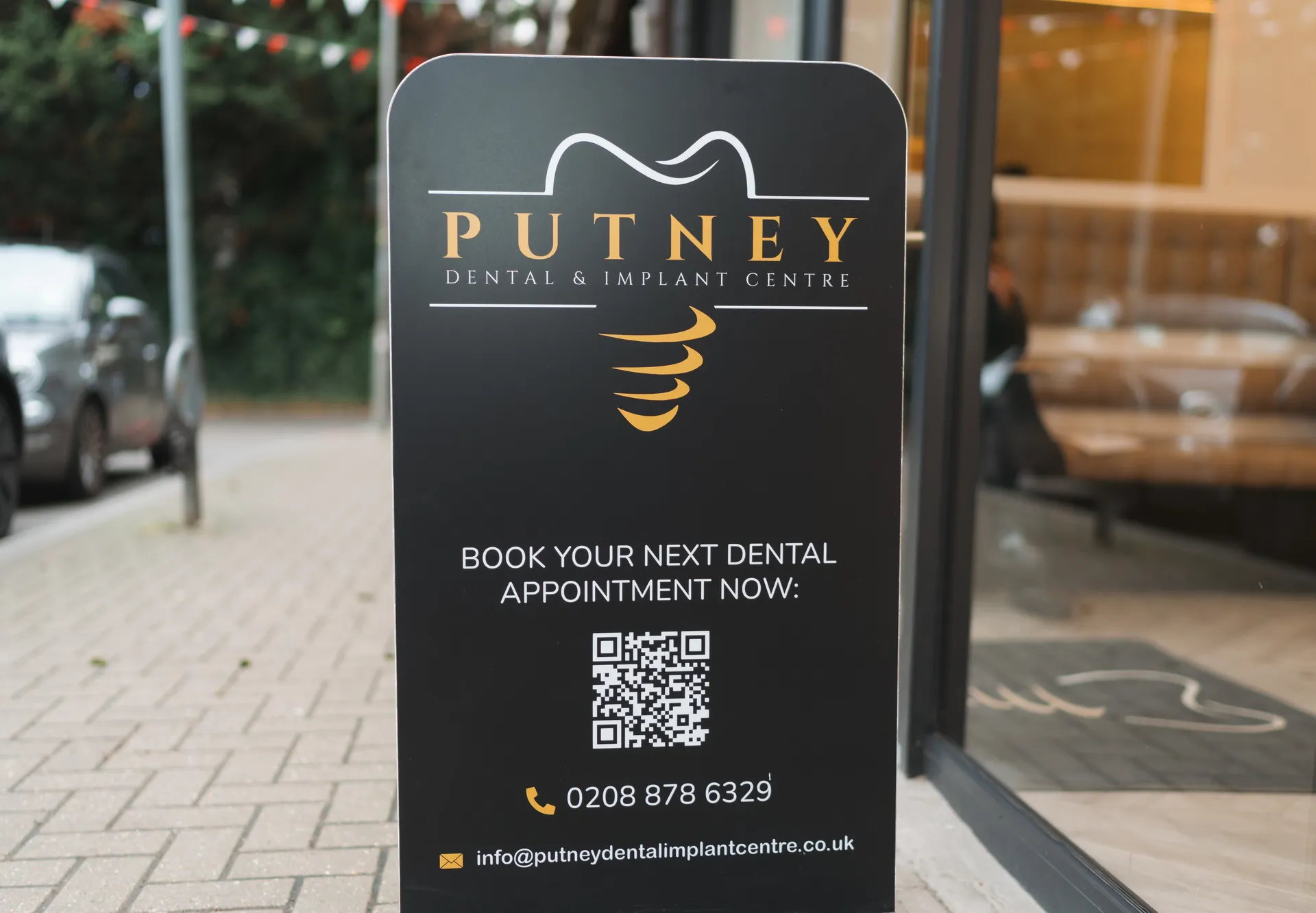 Black sidewalk sign for Putney Dental & Implant Centre with logo, QR code, phone number, and email for booking appointments.