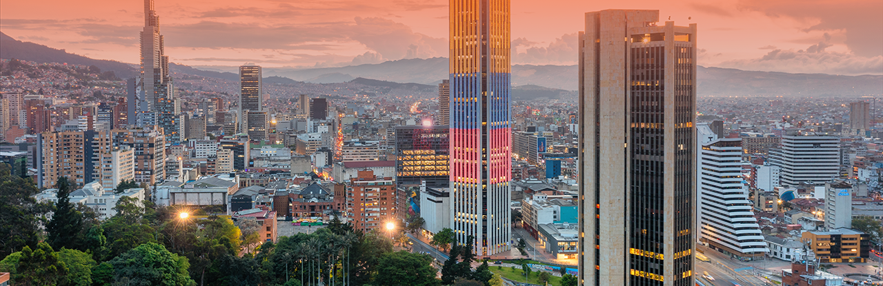 Cityscape of Bogotá, Colombia at sunset with tall buildings, including one with colorful vertical lights, and green trees in the foreground.