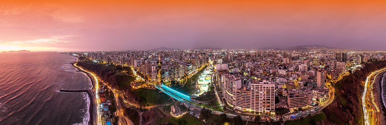 Panoramic aerial view of a coastal city at sunset with illuminated streets and buildings along cliffs by the ocean.