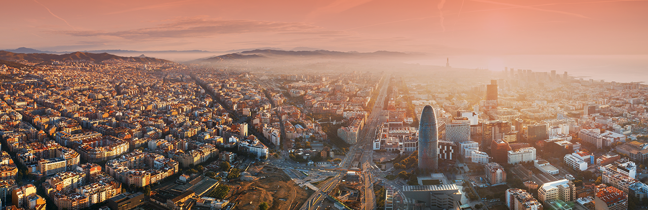 Aerial view of Barcelona cityscape at sunset featuring the distinctive Torre Glòries skyscraper and grid-pattern streets.