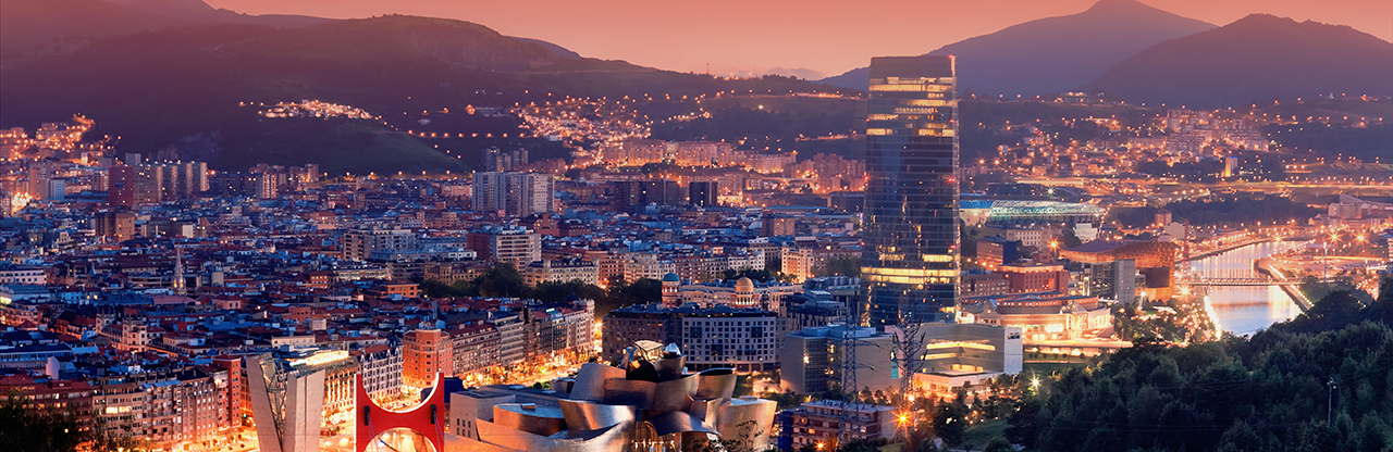 Panoramic cityscape at dusk with illuminated buildings, a modern museum with unique architecture, high-rise towers, and mountains in the background.