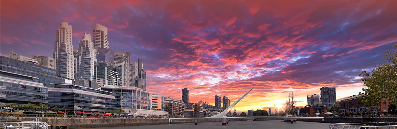 Modern city skyline with tall buildings, waterfront, and a white pedestrian bridge under a vibrant sunset sky.