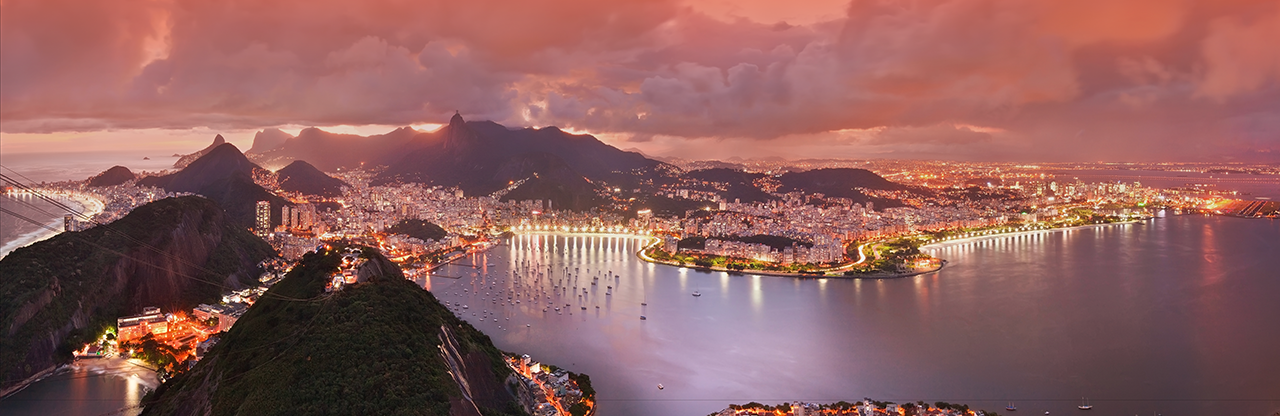 Aerial view of Rio de Janeiro at sunset with illuminated city, bay with boats, and surrounding mountains under a dramatic pink and orange sky.