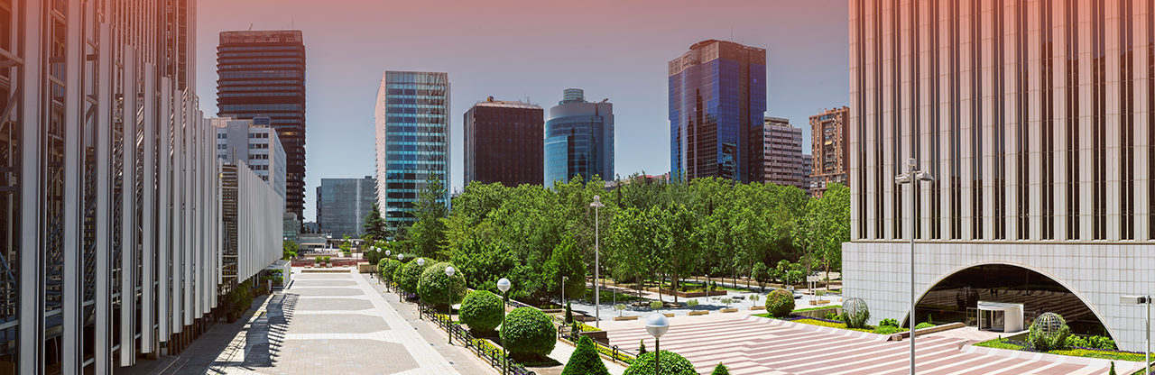 Urban plaza with symmetrical pathways flanked by trees, green shrubs, and modern office buildings under a clear sky.