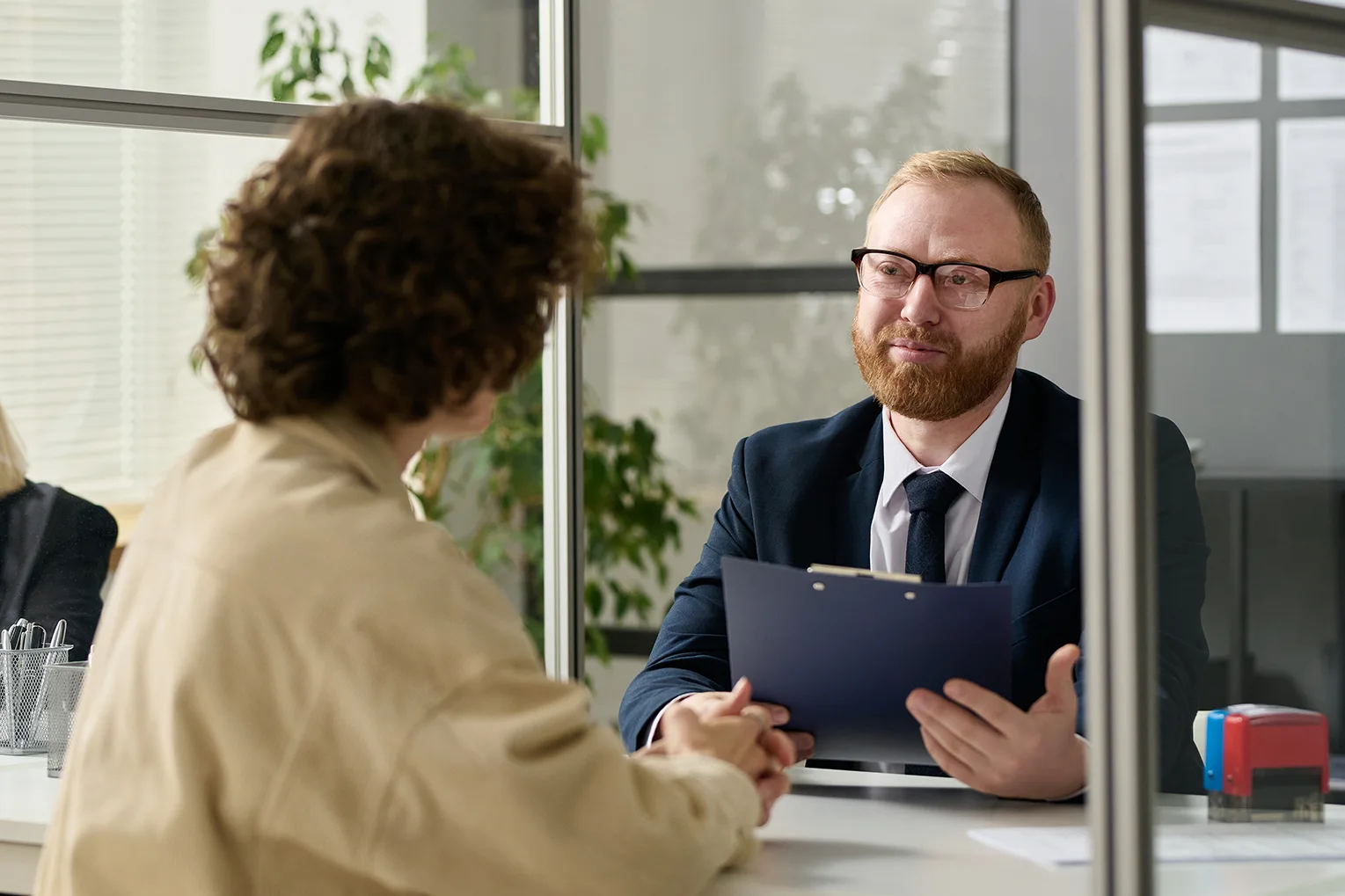 Professional man in a suit holding a clipboard while speaking with a client in a modern office setting.