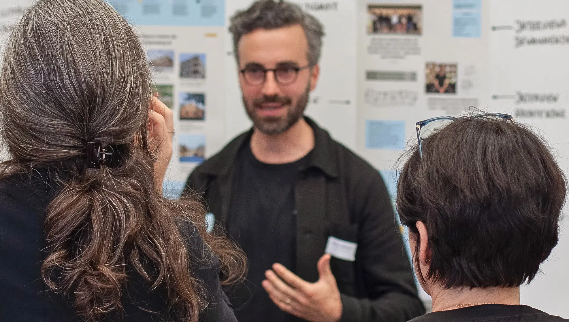 Ein Mann mit Brille spricht vor einer Wand mit Bildern und Texten, während zwei Frauen ihm zuhören.