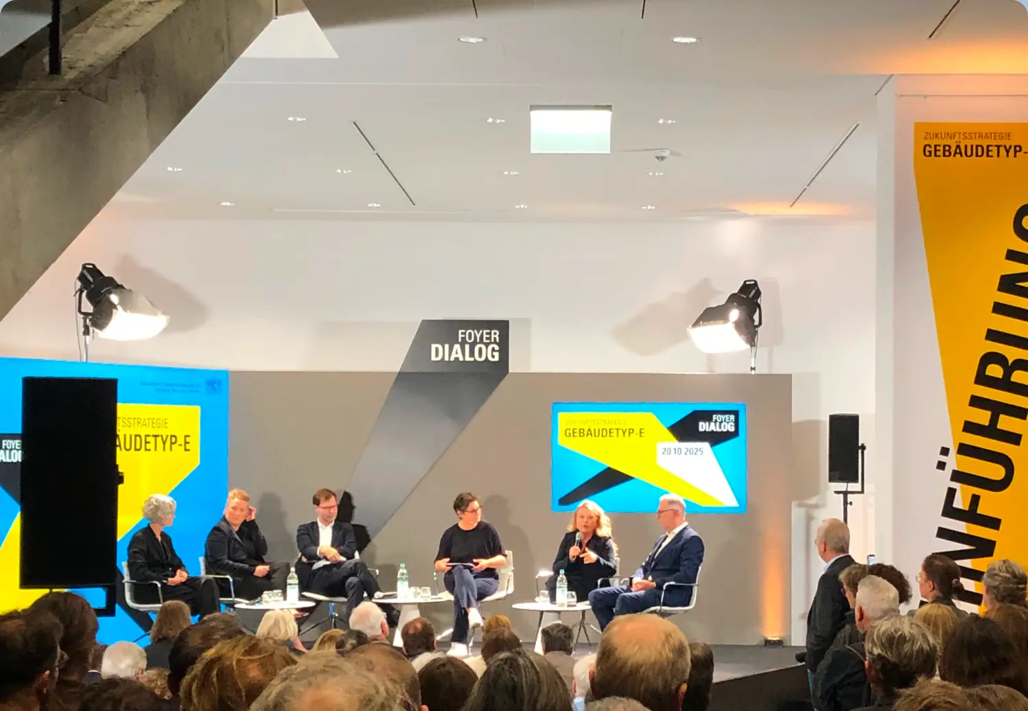 Podiumsdiskussion zum Gebäudetyp-E bei einer Architektur-Fachveranstaltung: Sechs Expertinnen und Experten sitzen auf der Bühne im Foyer Dialog und sprechen vor großem Publikum über Zukunftsstrategien im Bauwesen.