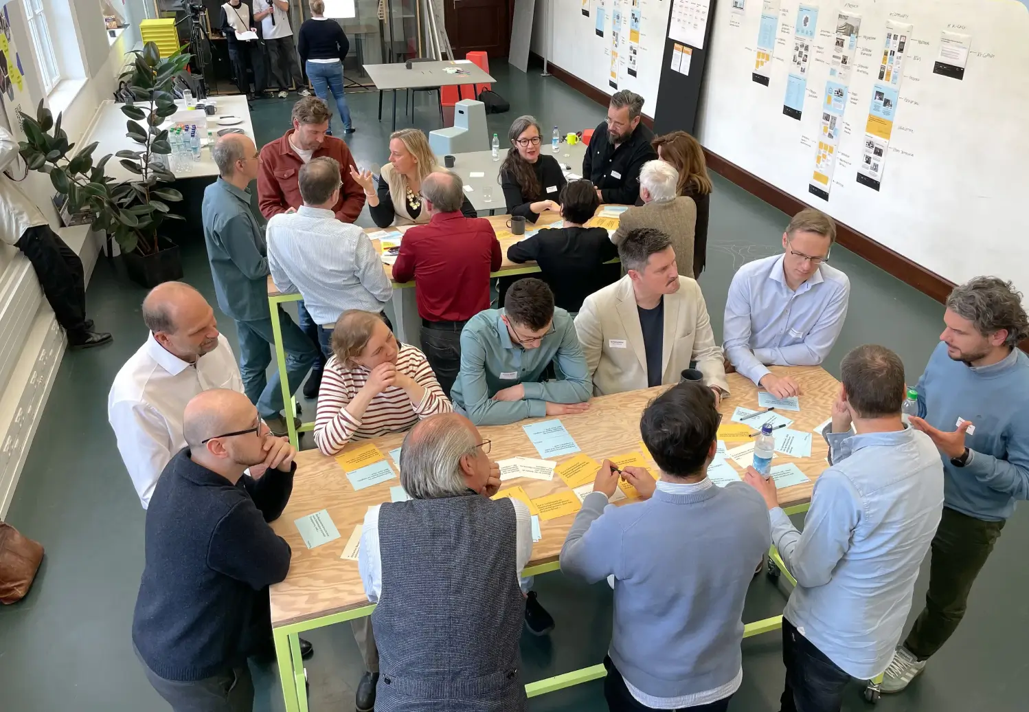 Workshop-Szene in einem offenen Kreativraum: Mehrere Gruppen von Fachleuten diskutieren intensiv an hohen Arbeitstischen über Konzeptkarten und Strategien, um gemeinsame Lösungen zu entwickeln.