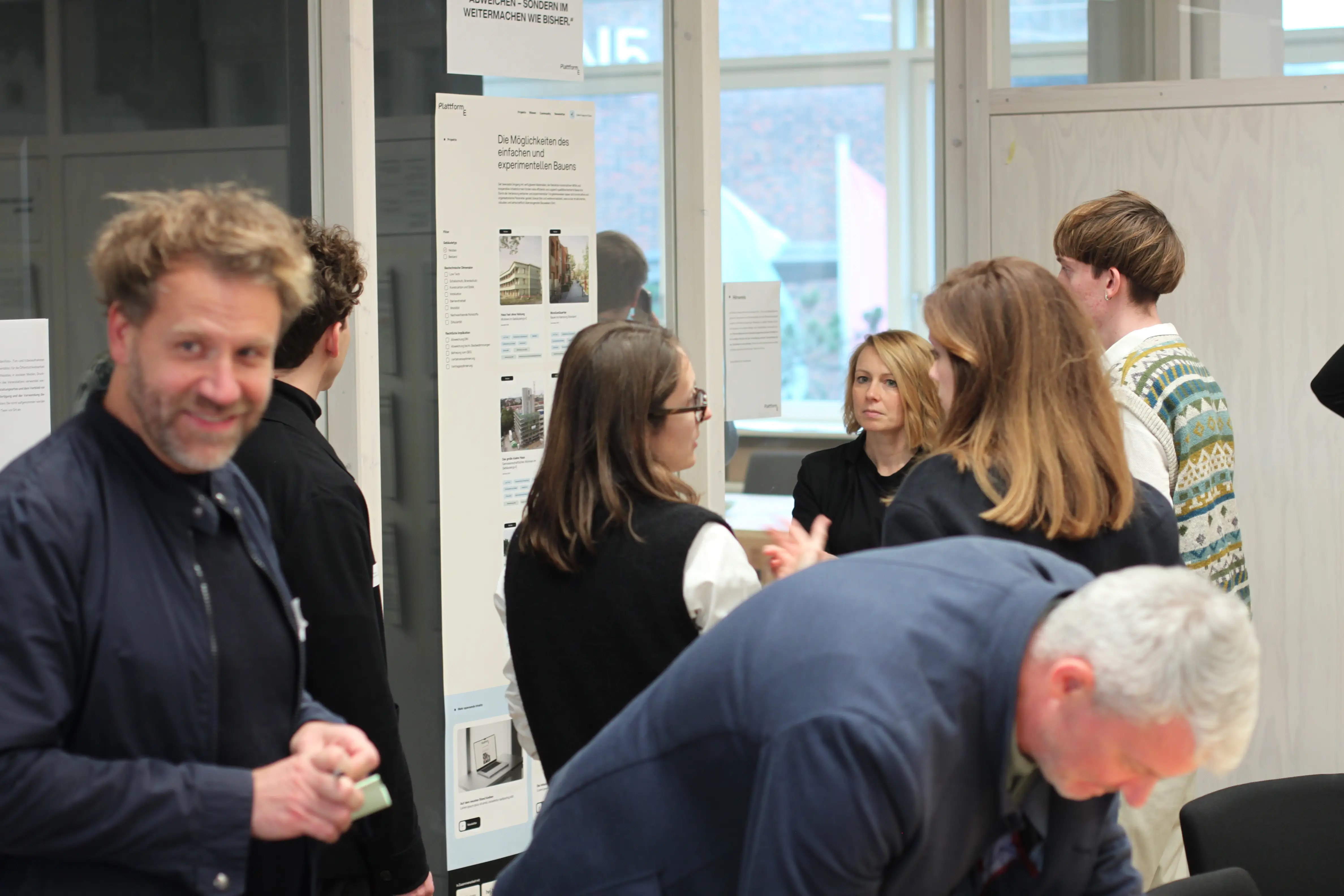Austausch während einer Architektur- und Planungsausstellung: Besucherinnen und Besucher diskutieren vor Infopostern über innovative Konzepte des experimentellen und einfachen Bauens.